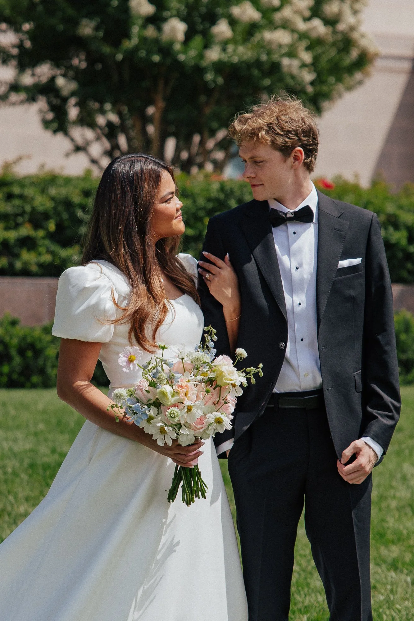 A bride and groom standing outdoors on a sunny day, looking at each other lovingly. The bride is holding a bouquet of white and pink flowers, wearing a white wedding dress with puffed sleeves. The groom is dressed in a black tuxedo with a bow tie. They are surrounded by greenery and trees.