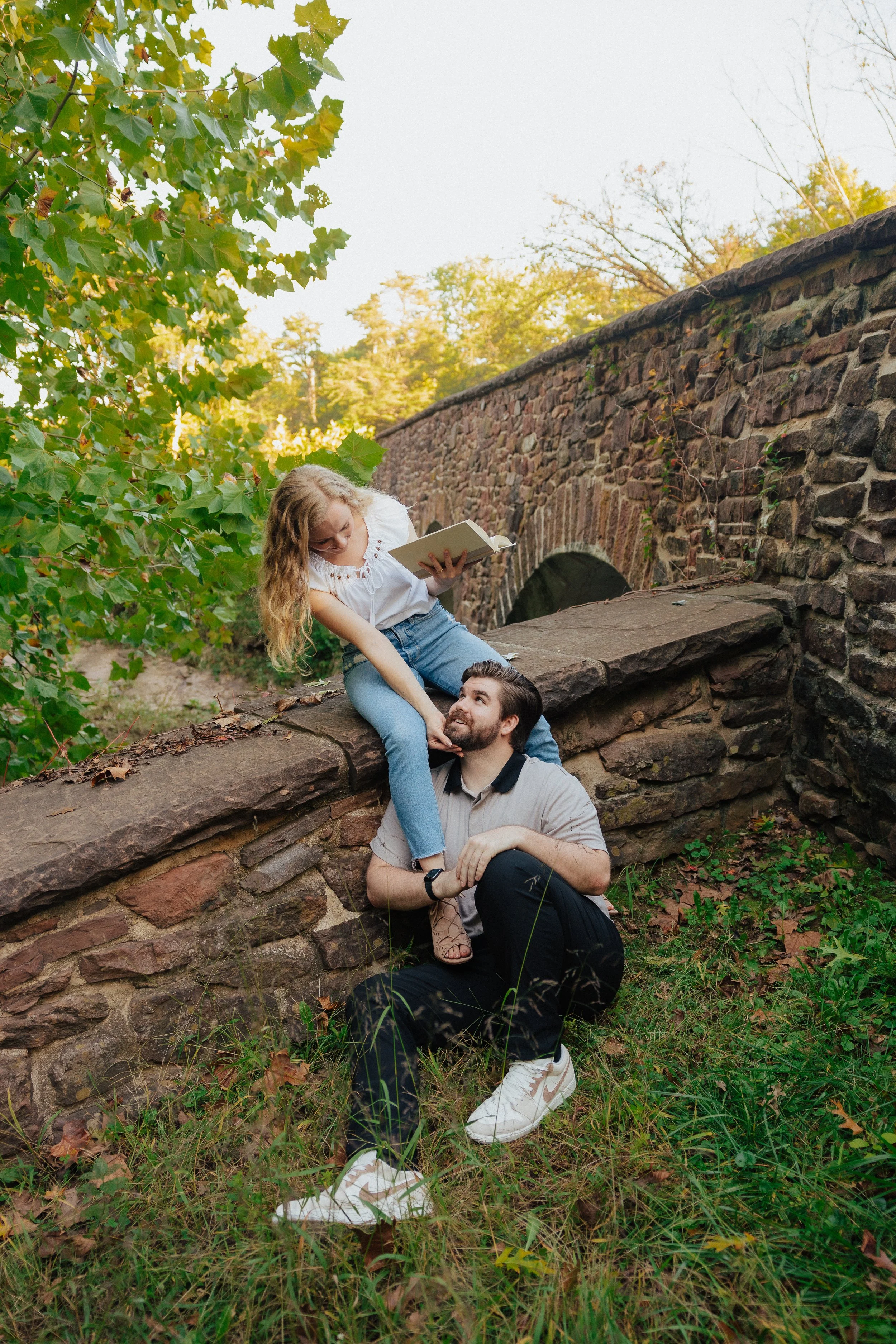 A young woman with long curly hair reading a book while sitting on a stone ledge, a man with a beard lying underneath her, holding her leg, in a park setting with trees and a stone bridge in the background.