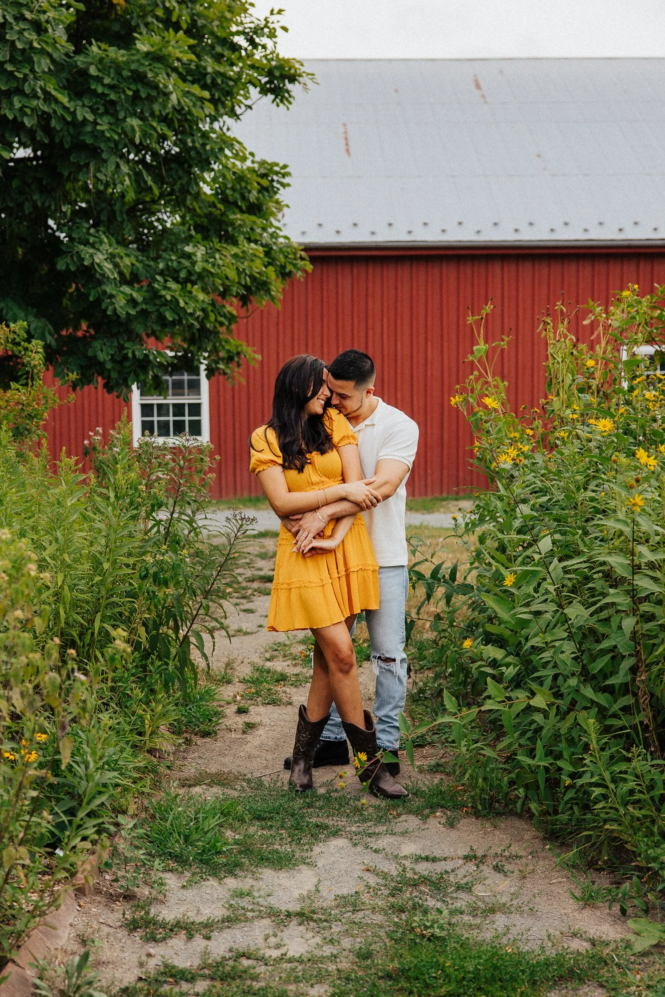 A smiling woman in a yellow dress and cowboy boots hugging a man in a white shirt and distressed jeans on a farm path, surrounded by green plants and a red barn in the background.