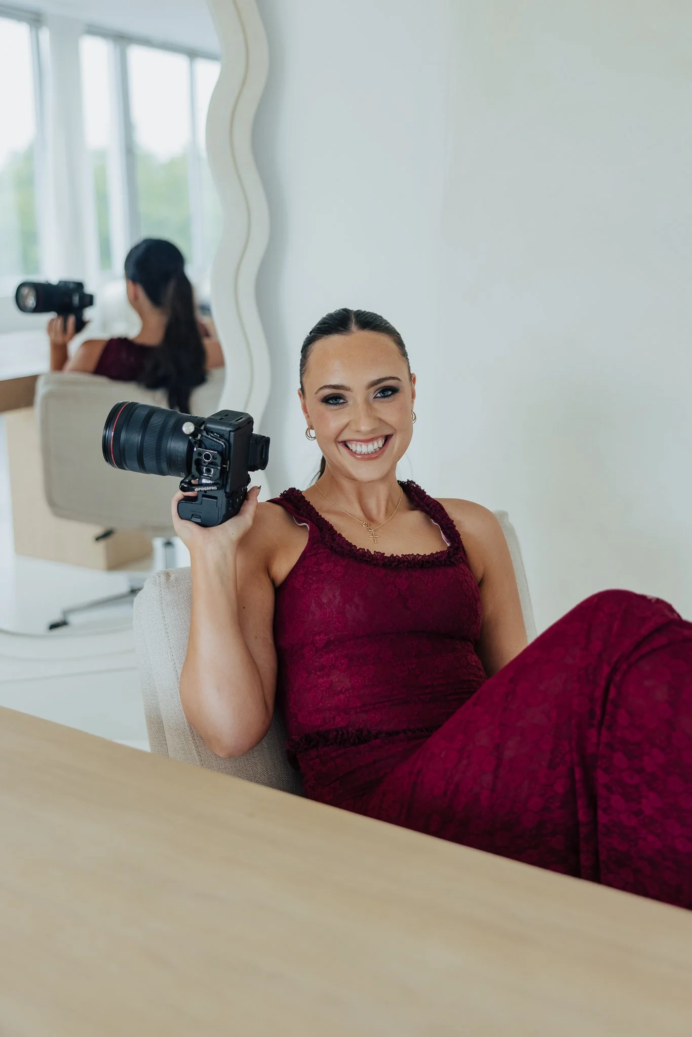 A woman in a burgundy dress smiling and holding a camera, sitting in a room with a large mirror and a white wall.