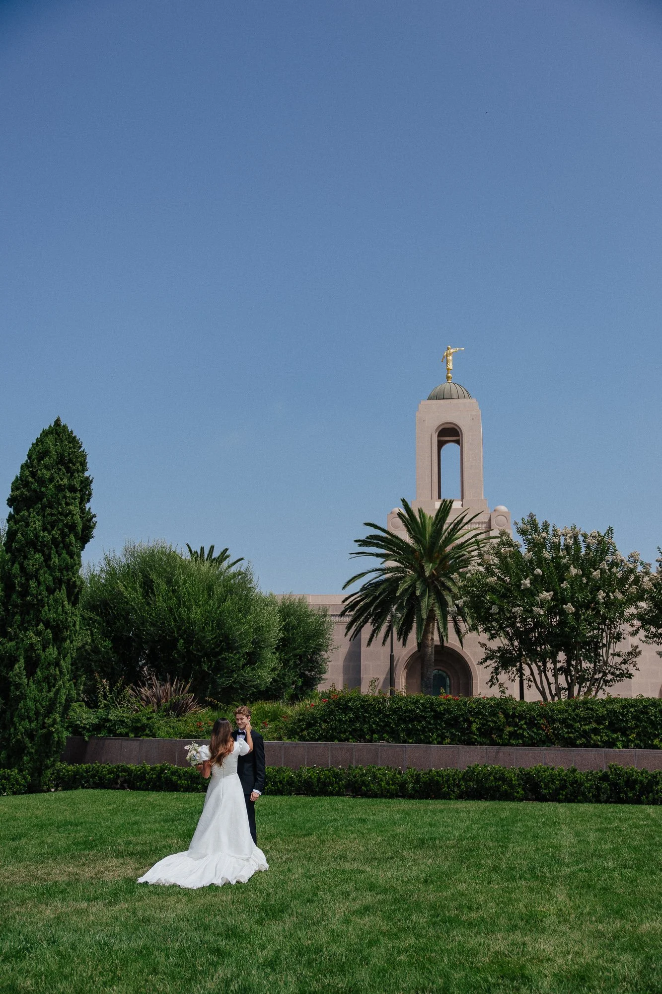 A bride and groom standing on a lush green lawn in front of a historical building with a tower featuring a golden sculpture on top. The couple is celebrating their wedding, with the bride in a white gown holding a bouquet. The scene is set in a landscaped garden with various trees and shrubs under a clear sky.