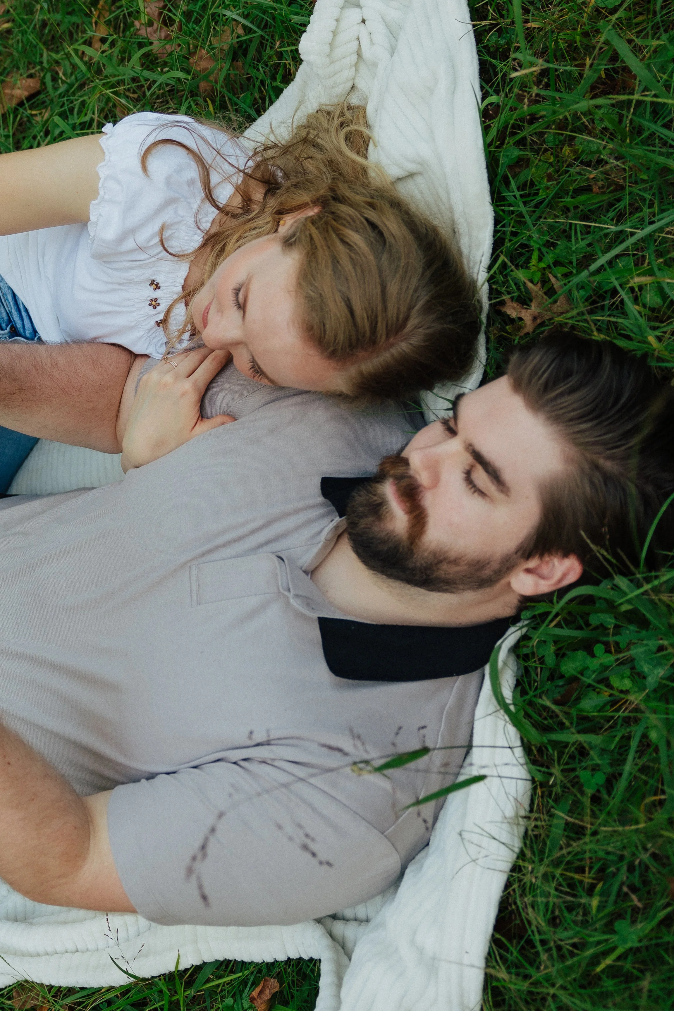 A woman with wavy blonde hair lying on a white blanket in green grass, resting her head on a man with a beard, both appearing relaxed with eyes closed.