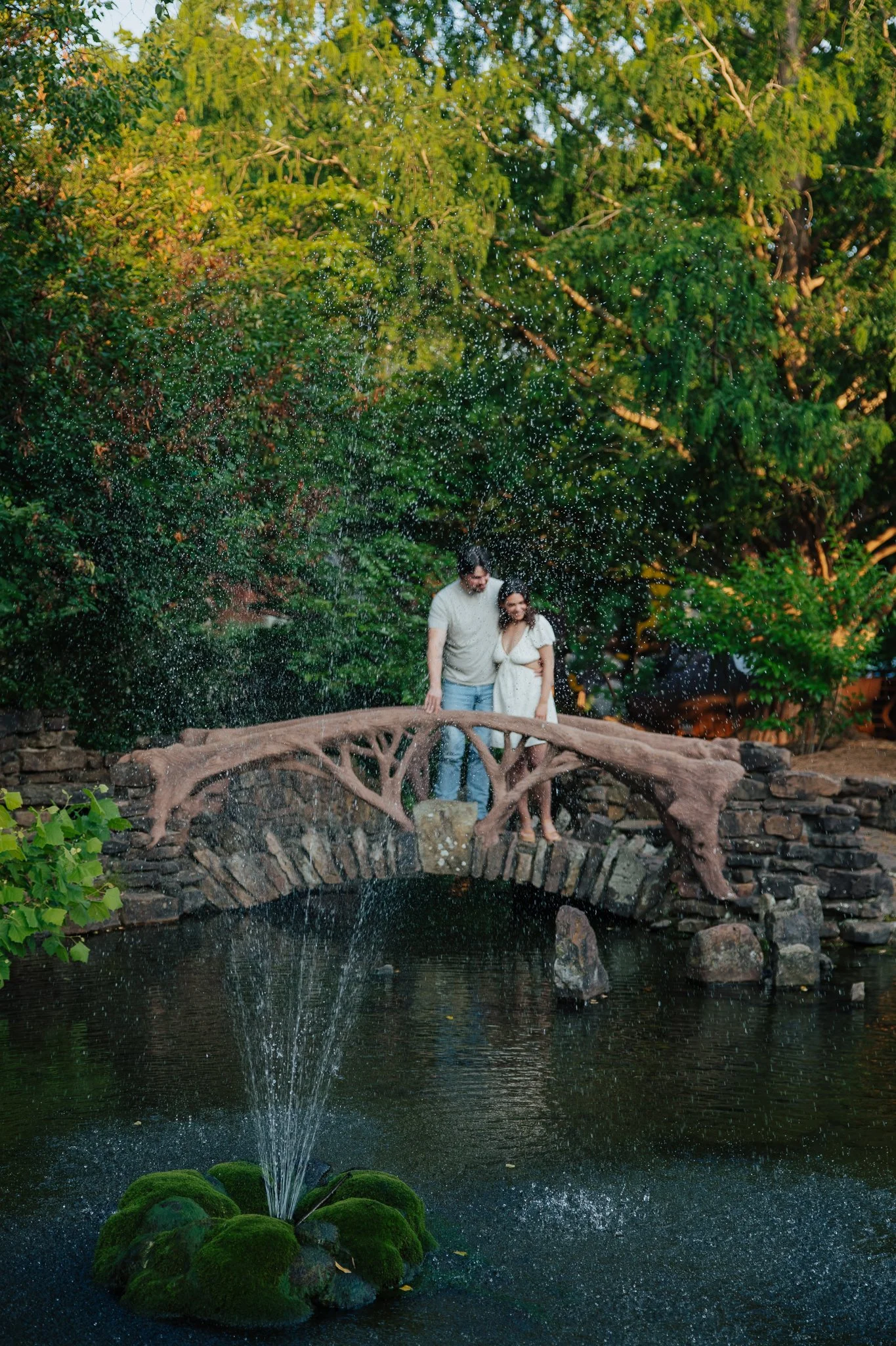 A couple standing on a small stone and wood bridge over a pond with a fountain and moss-covered rocks, surrounded by lush green trees and foliage.