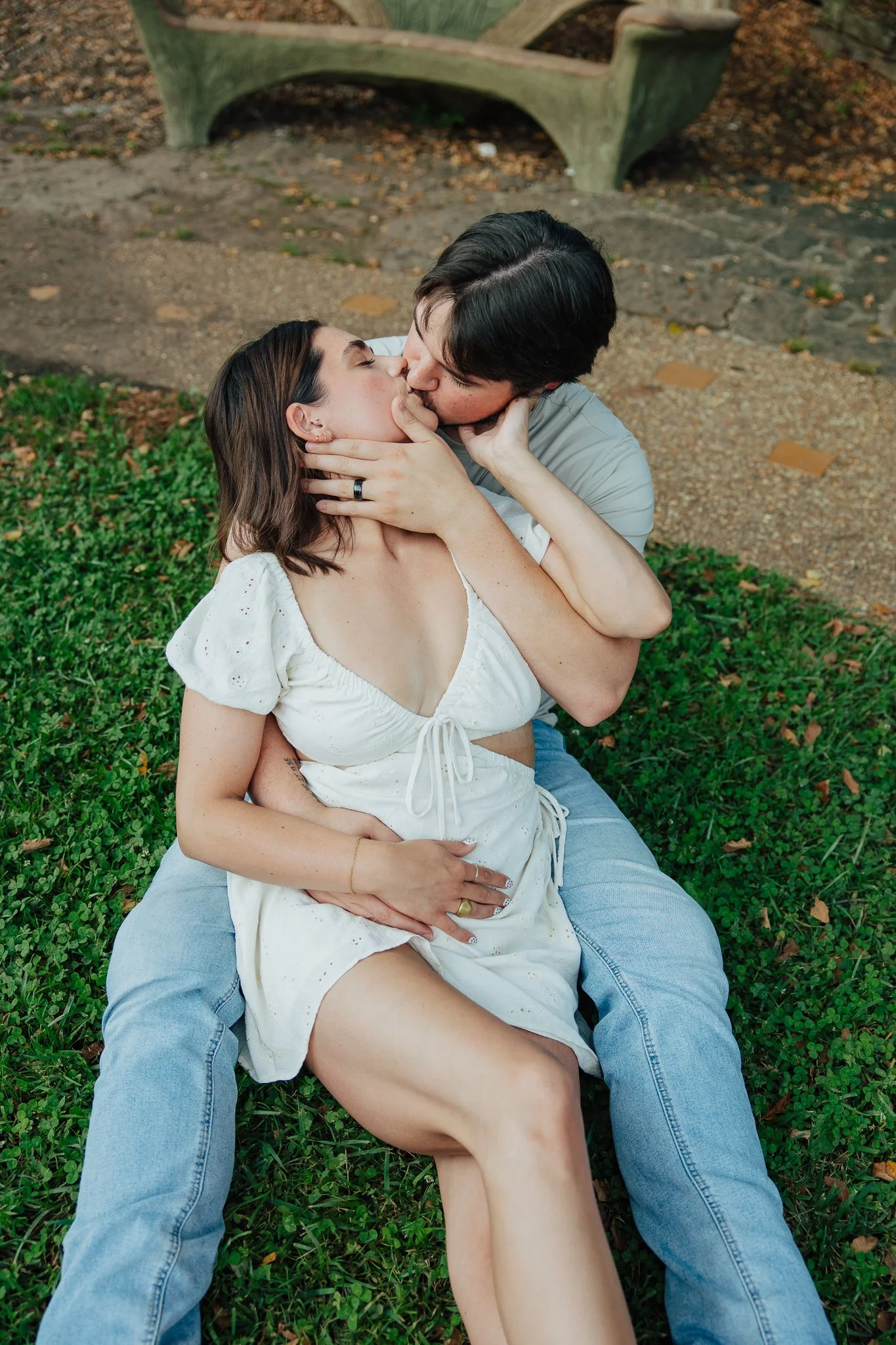 A couple is kissing while lying on the grass in a park, with a wooden bench in the background.