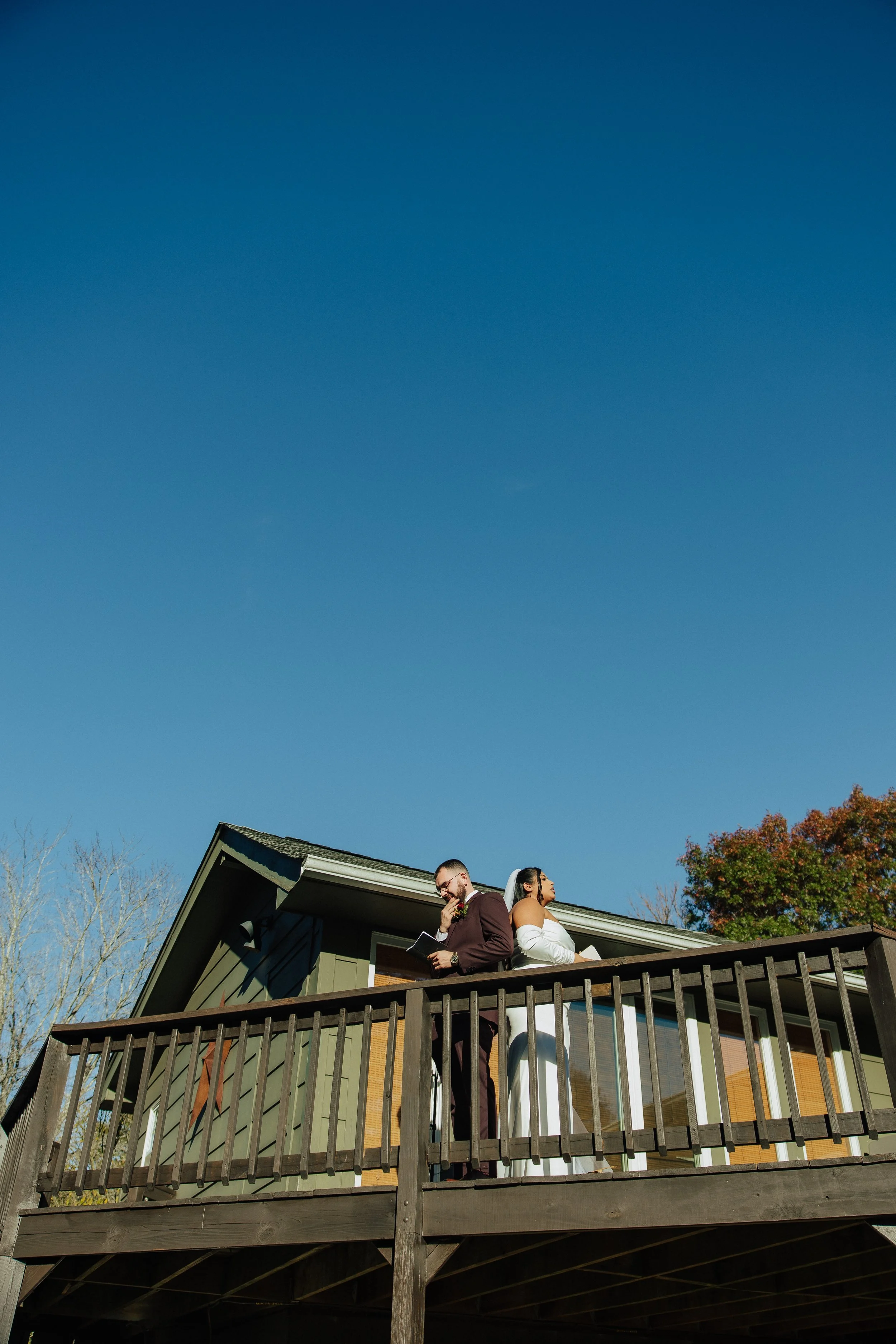 A man and woman dressed in wedding attire standing on a balcony of a house, with a clear blue sky in the background and trees around.