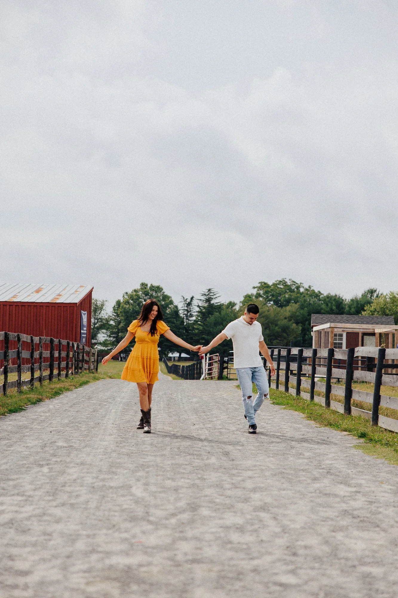 A young woman in a yellow dress and cowboy boots holding hands with a young man in a white shirt and ripped jeans walking down a gravel path in a rural area with farm buildings and trees in the background.