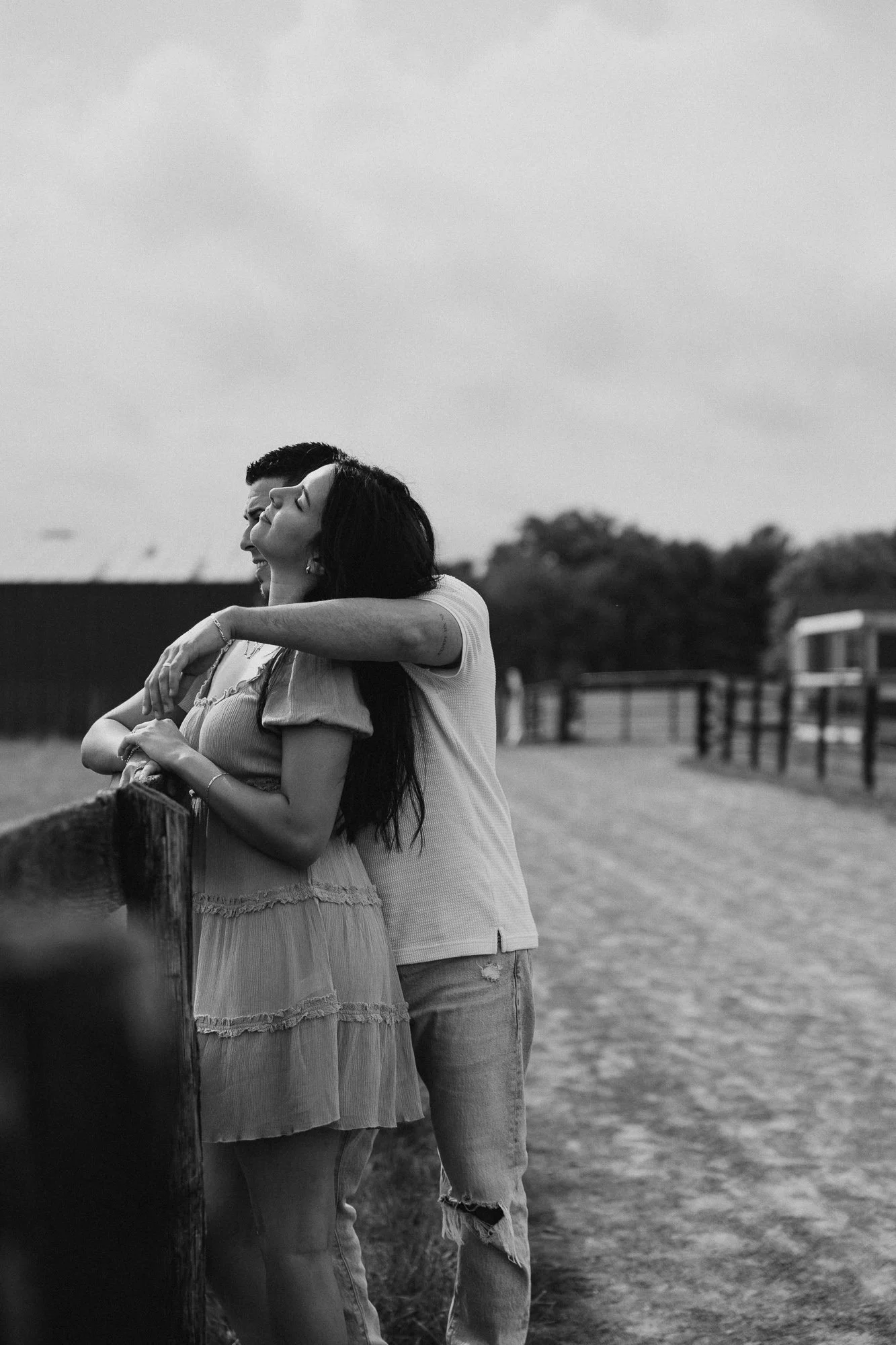 A black and white photo of a woman and a man embracing outdoors on a cloudy day, with a rural landscape in the background.