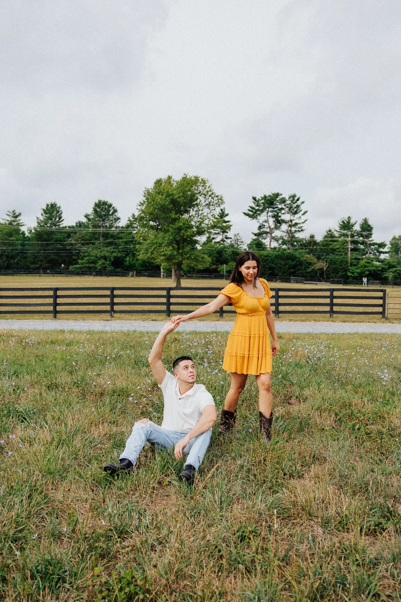 A woman in a yellow dress standing in a field, holding the hand of a man who is sitting on the ground, with a background of trees and cloudy sky.