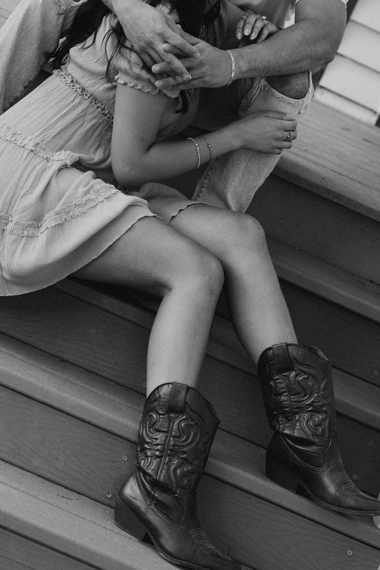 Close-up black and white photo of a woman and man sitting close together on a wooden bench, with the woman's arm around the man's leg and their hands intertwined. The woman is wearing a ruffled skirt, bracelets, and rings, and the man is wearing ripp