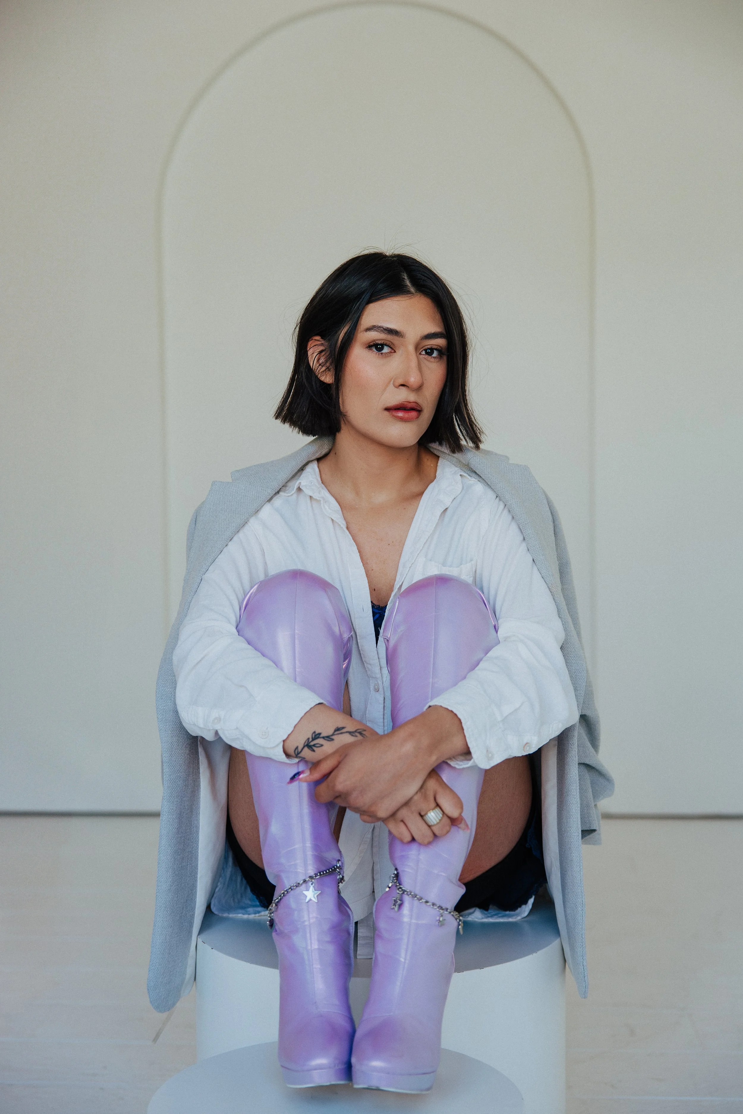 A woman with dark hair and a white shirt sitting on a stool with purple boots, arms crossed, in front of a plain beige and white background.
