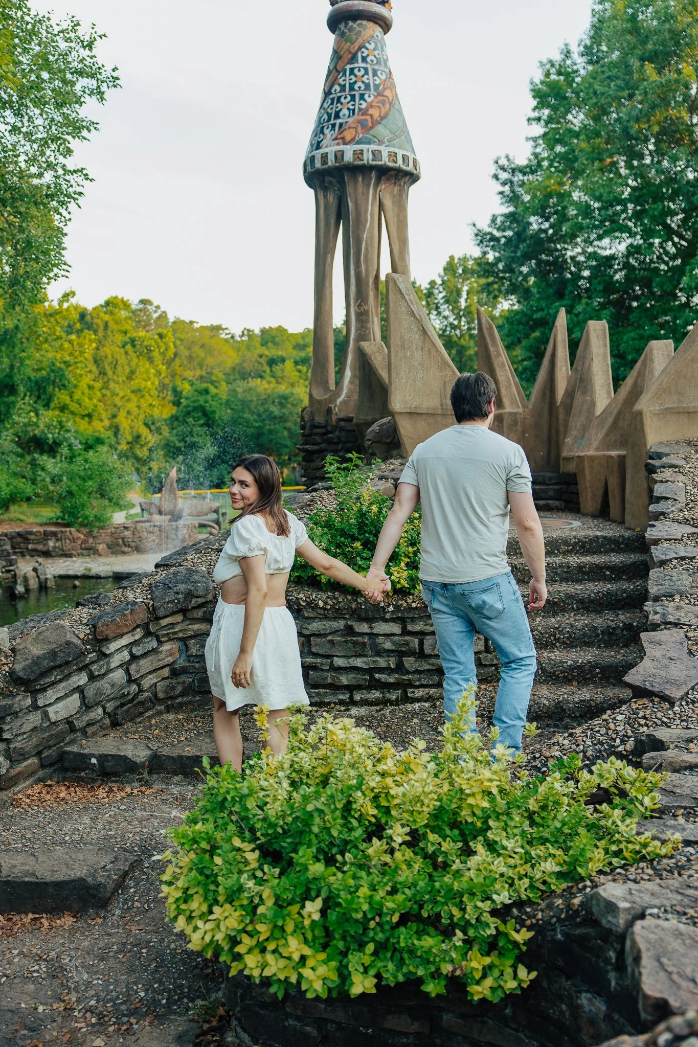 A couple holding hands walking through a garden with a stone wall and greenery, with a large outdoor sculpture of a giraffe in the background.