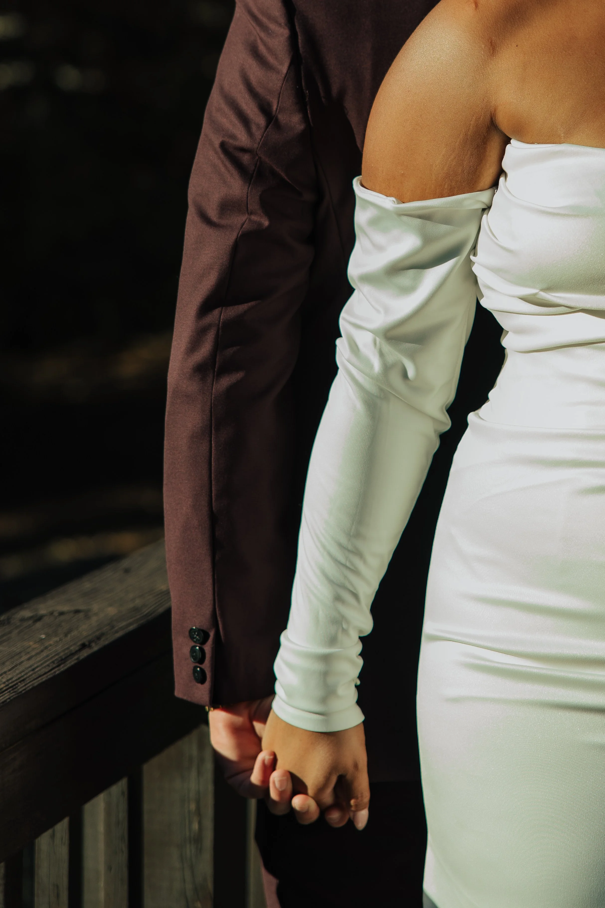 A couple holding hands, with the woman in a white off-the-shoulder dress and the man in a dark suit, standing outdoors on a wooden bridge.