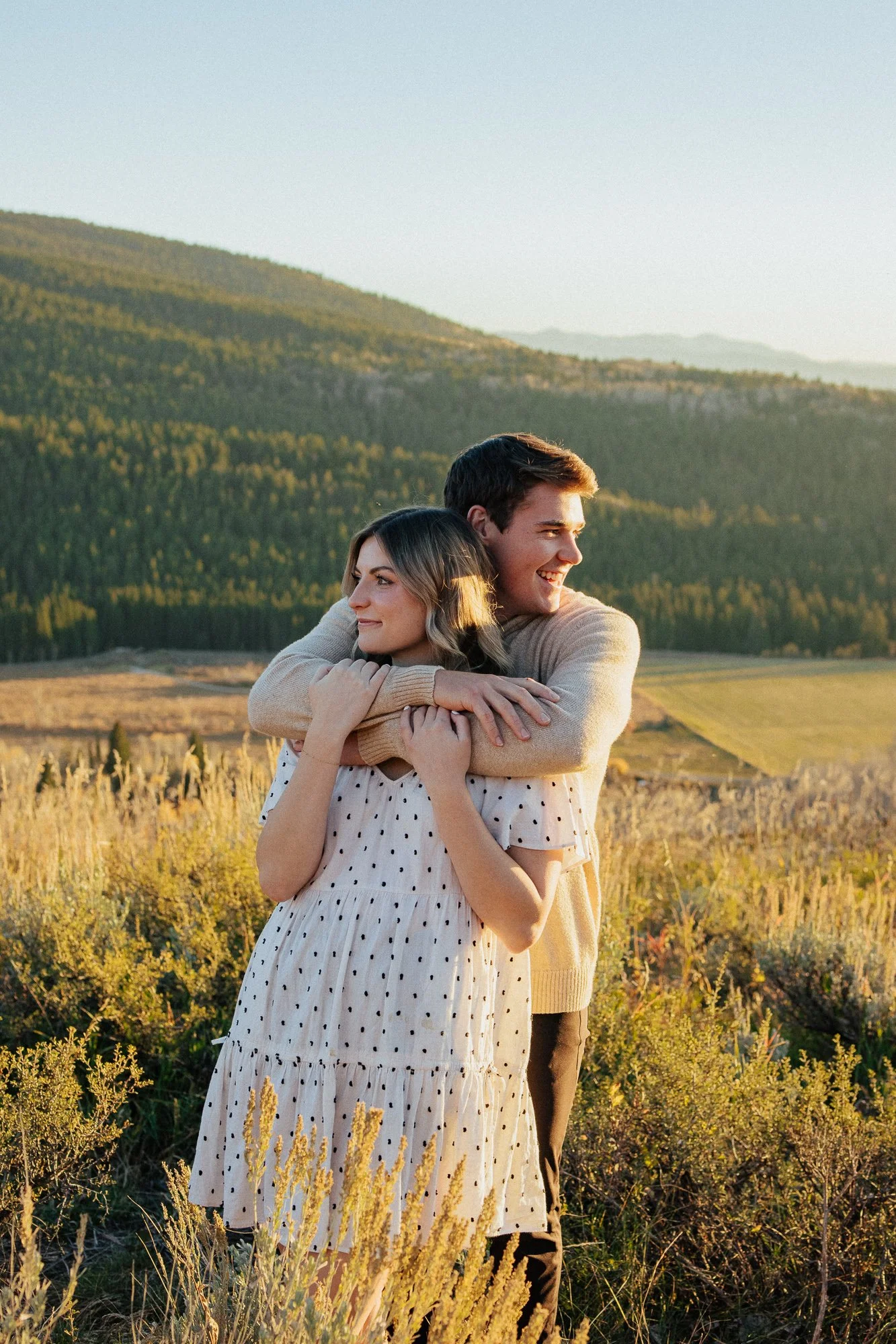 A young couple embracing in a field of tall grass and wildflowers with a lush green mountain range and a clear sky in the background, taken during golden hour.