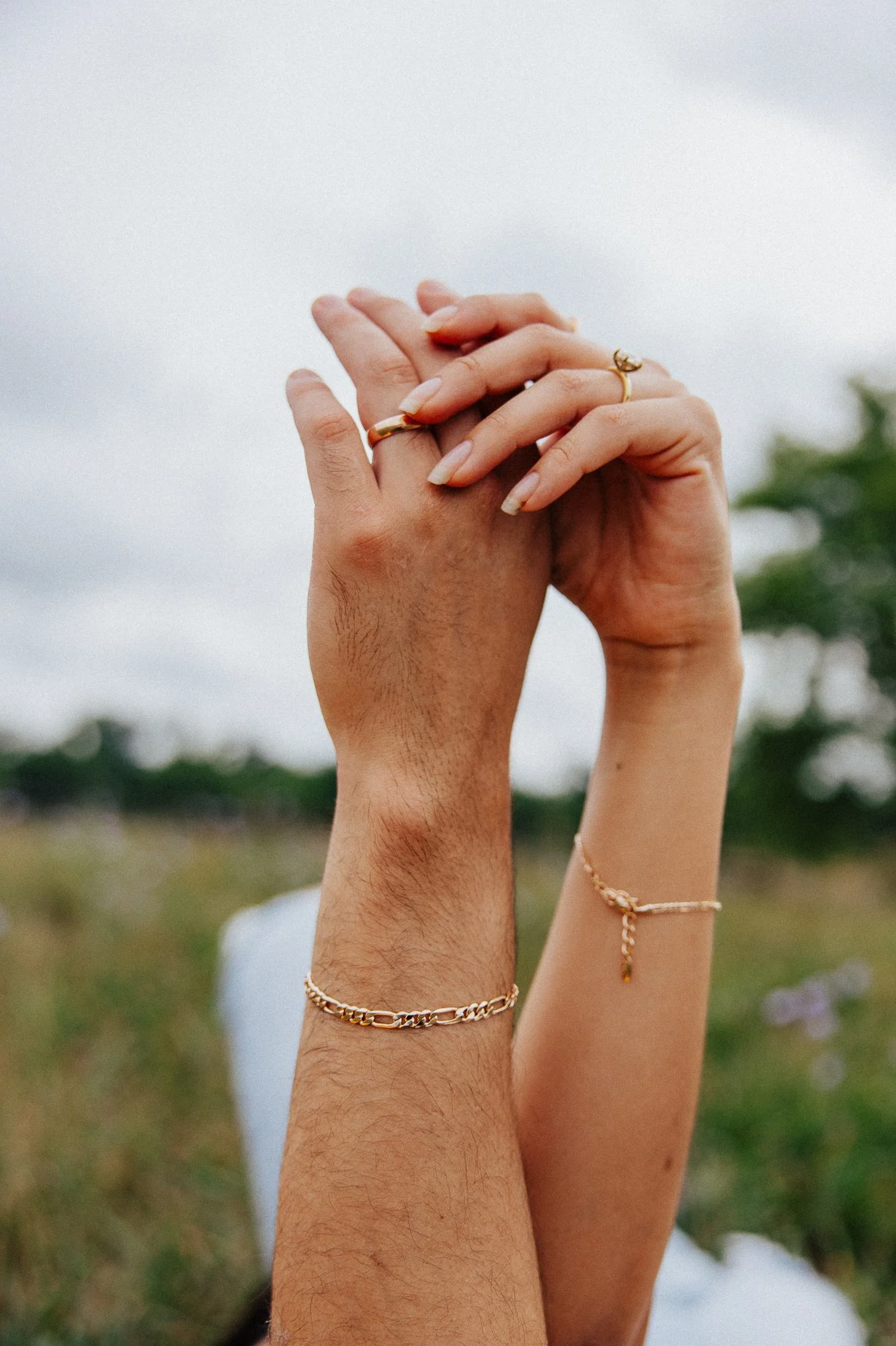 Two hands, one male and one female, intertwined with fingers touching against a cloudy sky with greenery in the background. The female hand is adorned with rings and bracelets.