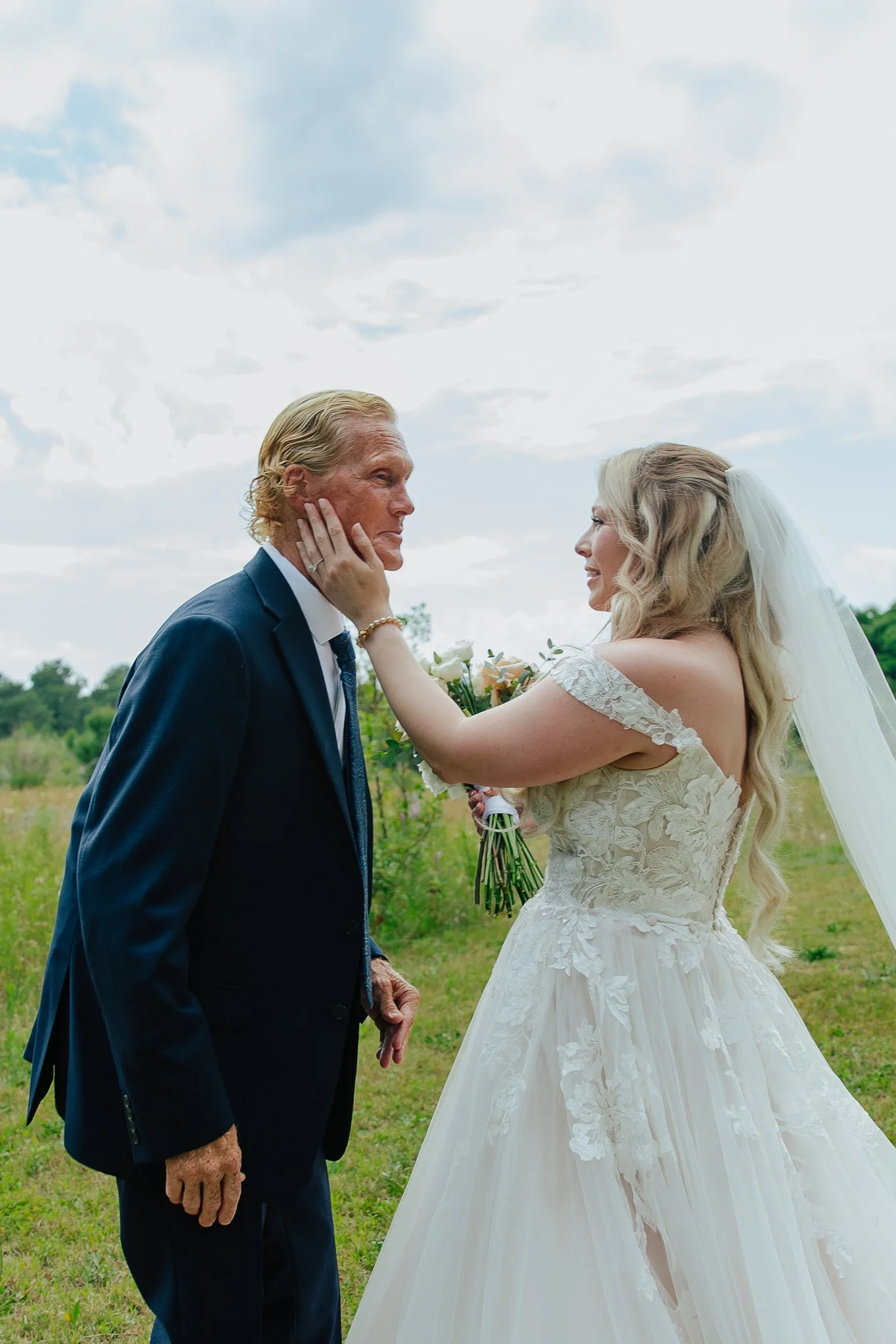 A bride with long blonde hair in a lace wedding dress touching the face of an older groom in a navy suit, outdoors on a grassy field with a cloudy sky.