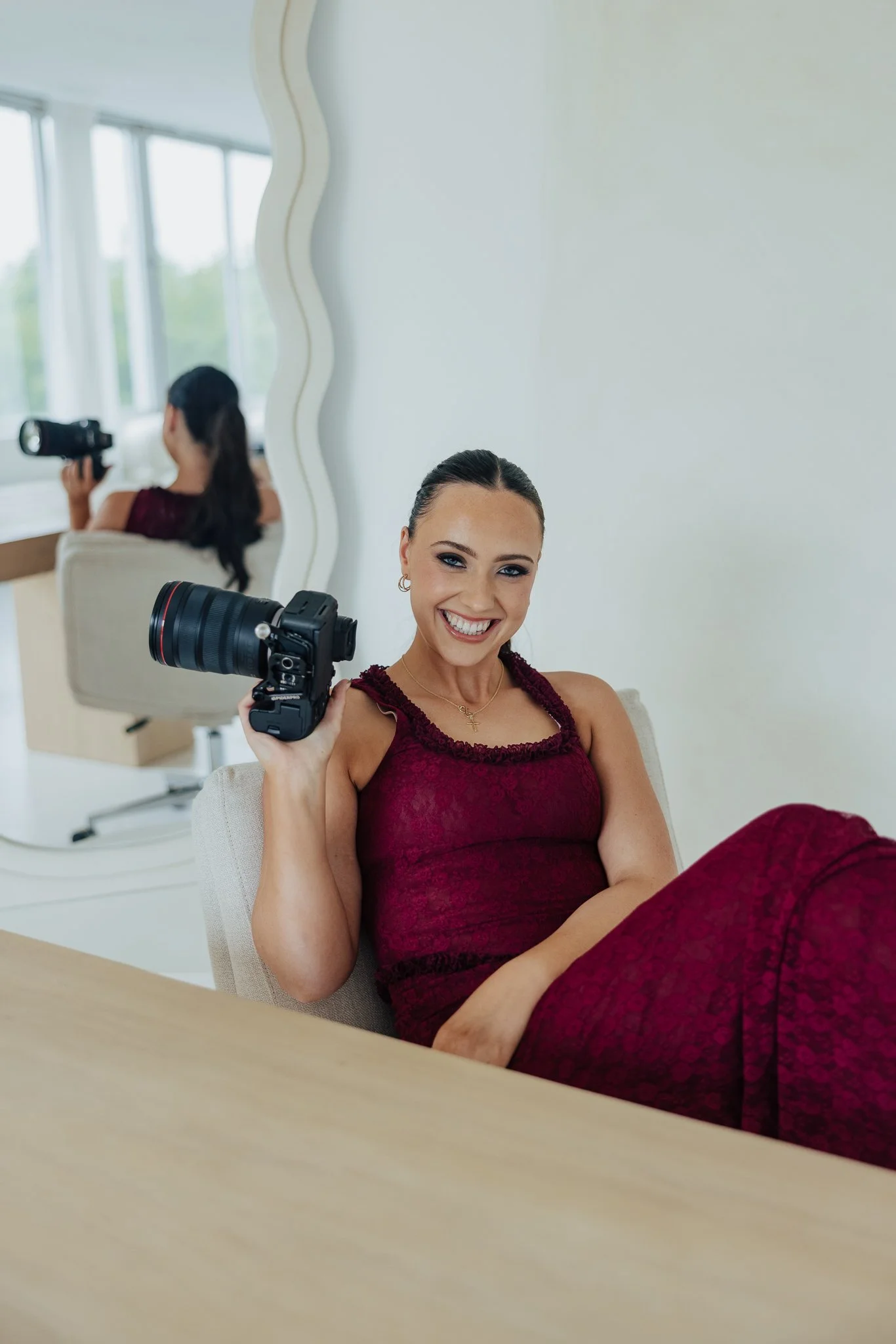 A woman in a burgundy dress smiling and holding a large camera in a well-lit room with a mirror and a woman with a camera in the background.