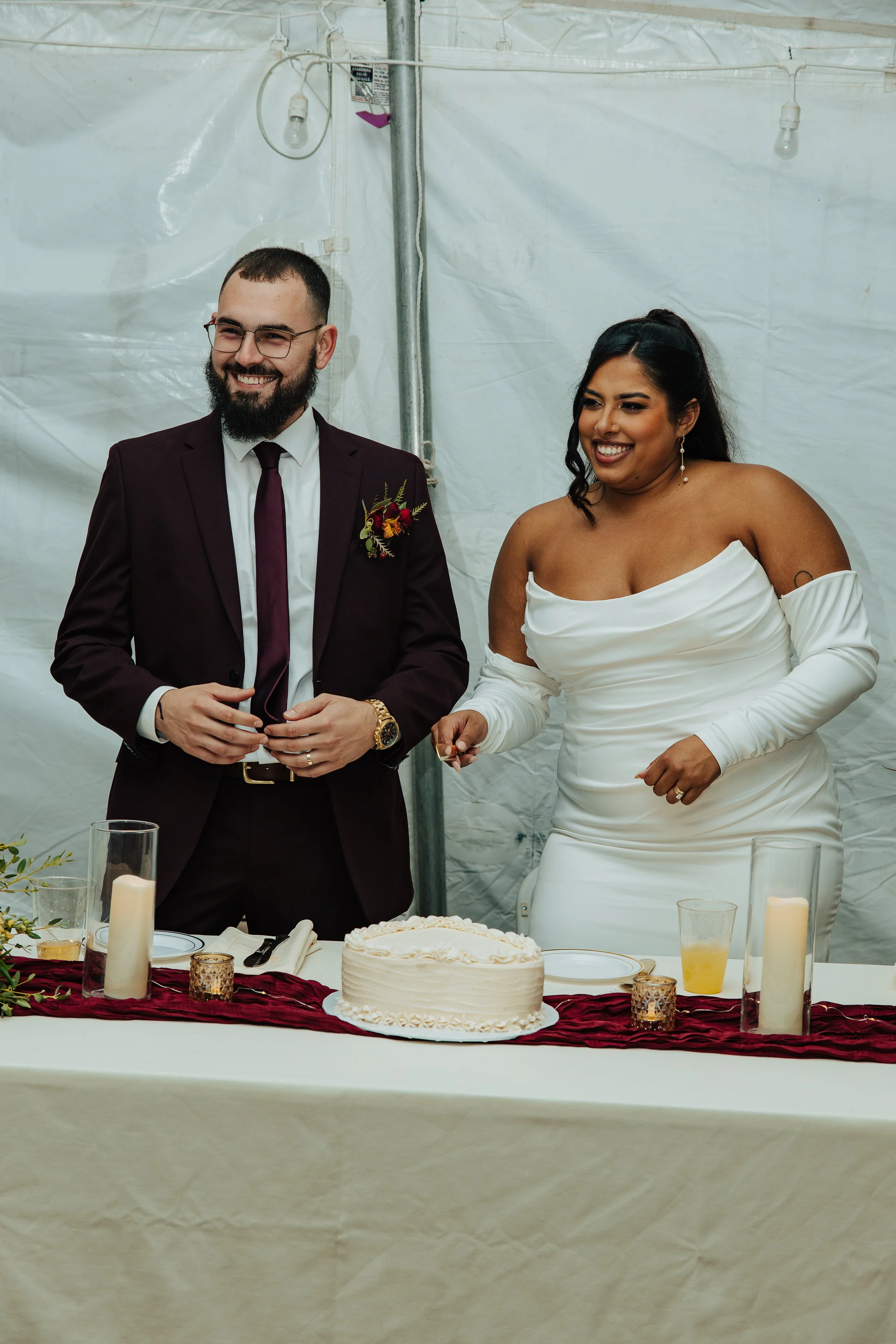 A man in a dark suit and a woman in a white wedding dress smiling at a wedding reception. There are candles and a wedding cake on the table.