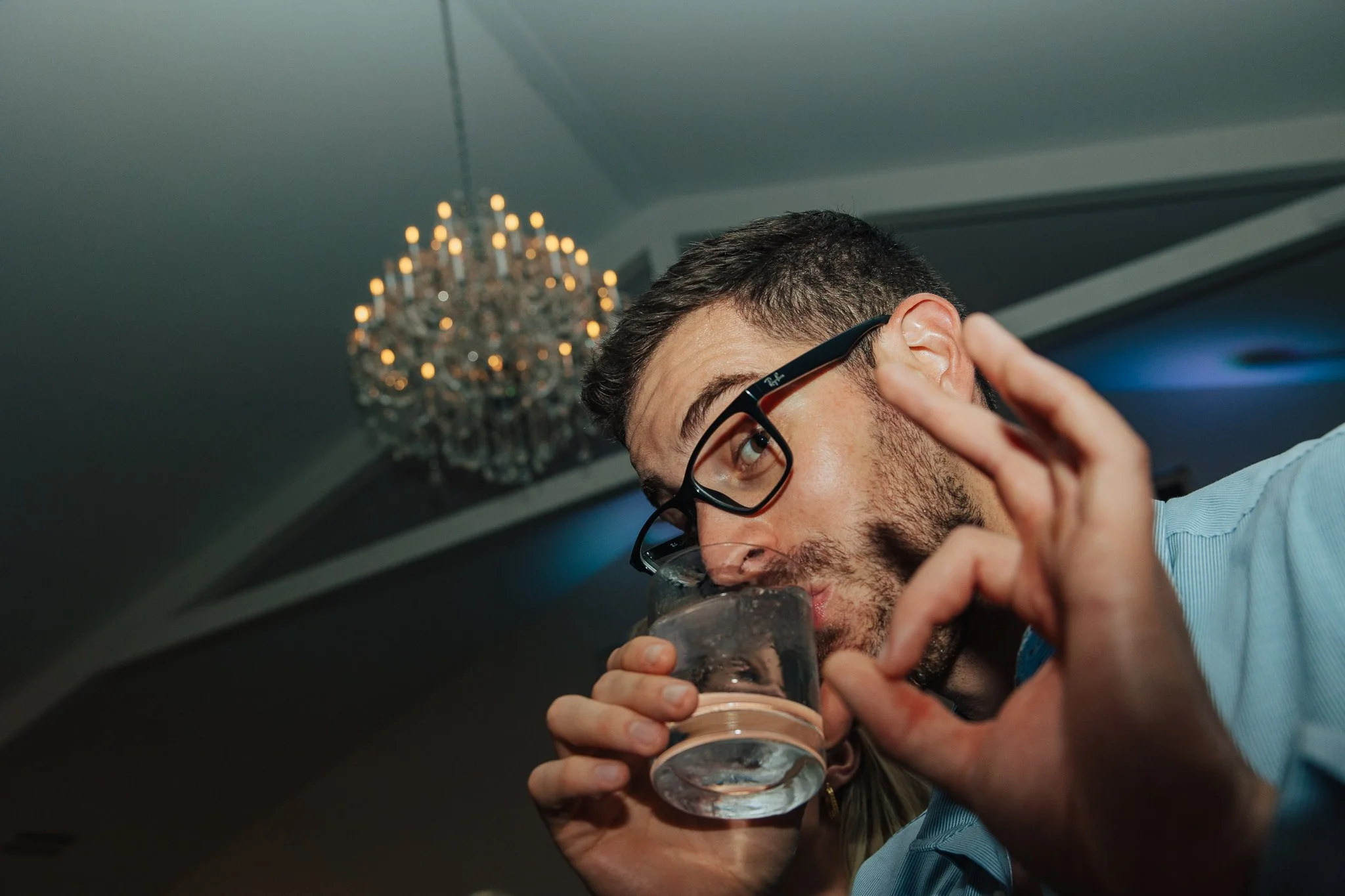 A man wearing glasses is drinking from a glass of water. There is a chandelier hanging from the ceiling in the background.