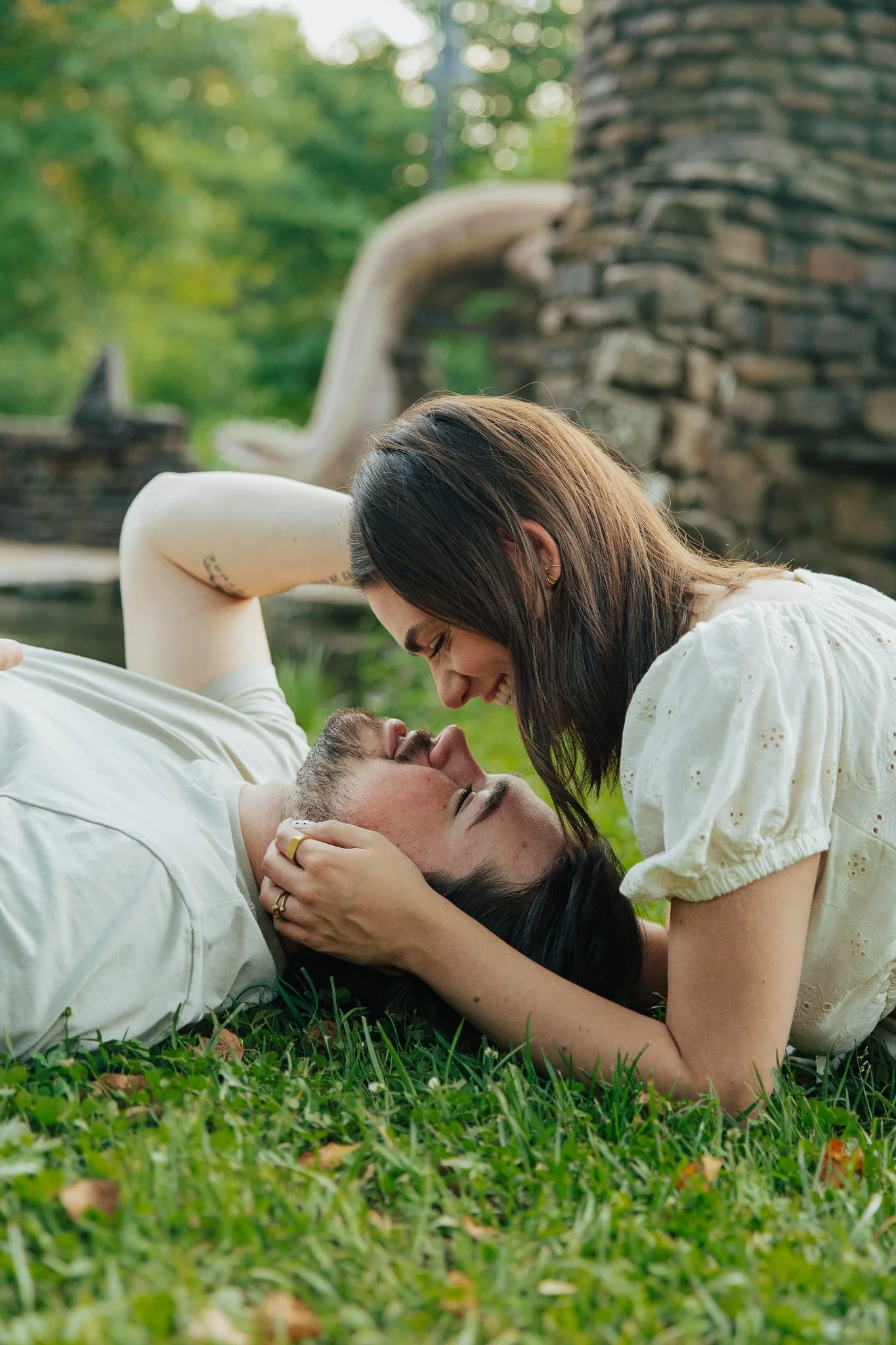 A couple lying on the grass outside, with the woman leaning over the man, smiling and holding his face, in a park or garden setting.
