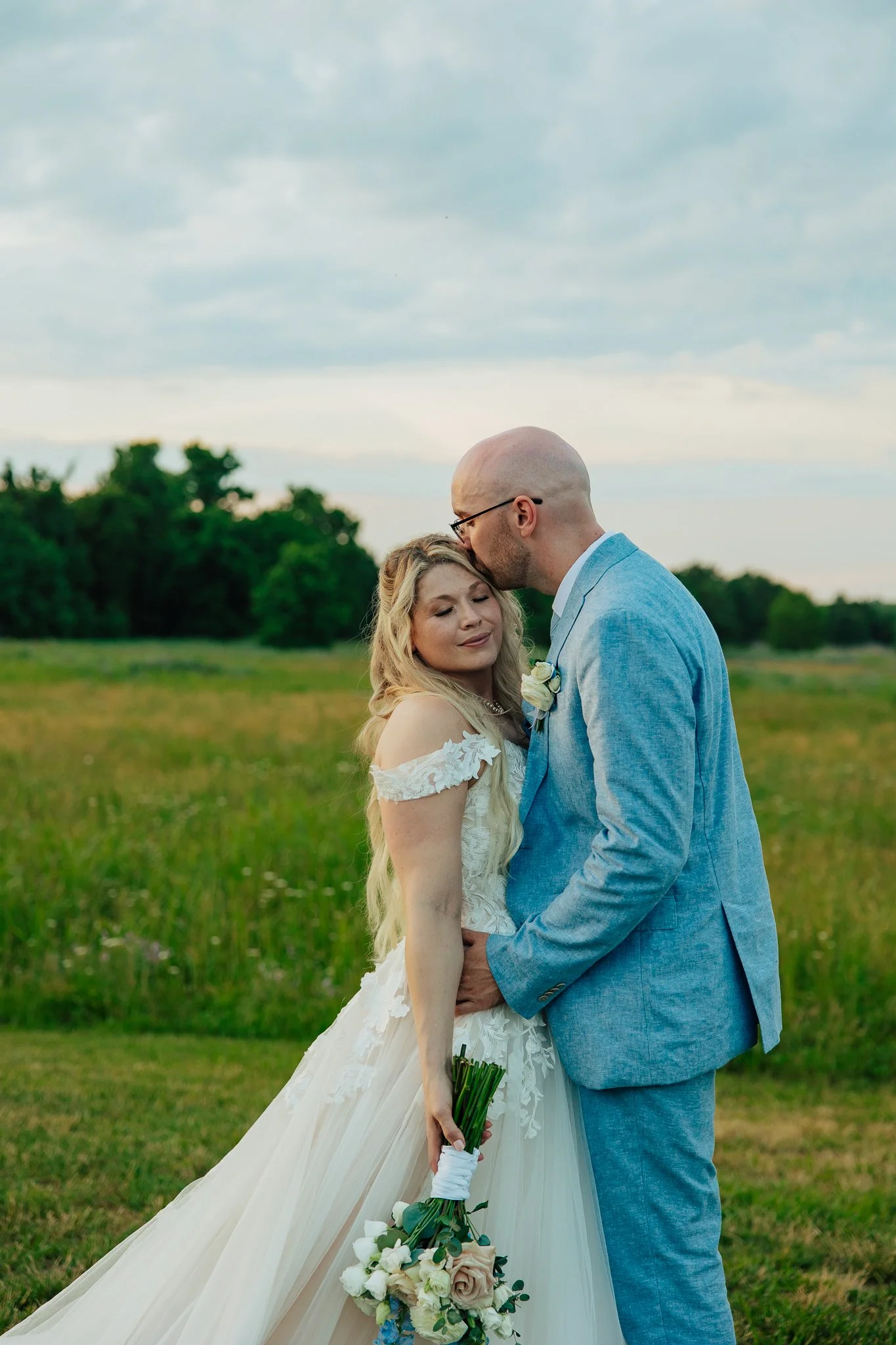 A bride in a white wedding dress holding a bouquet of white and pale pink roses, standing with a groom in a light blue suit outdoors in a green field with trees and a cloudy sky in the background. The groom is kissing the bride's forehead.