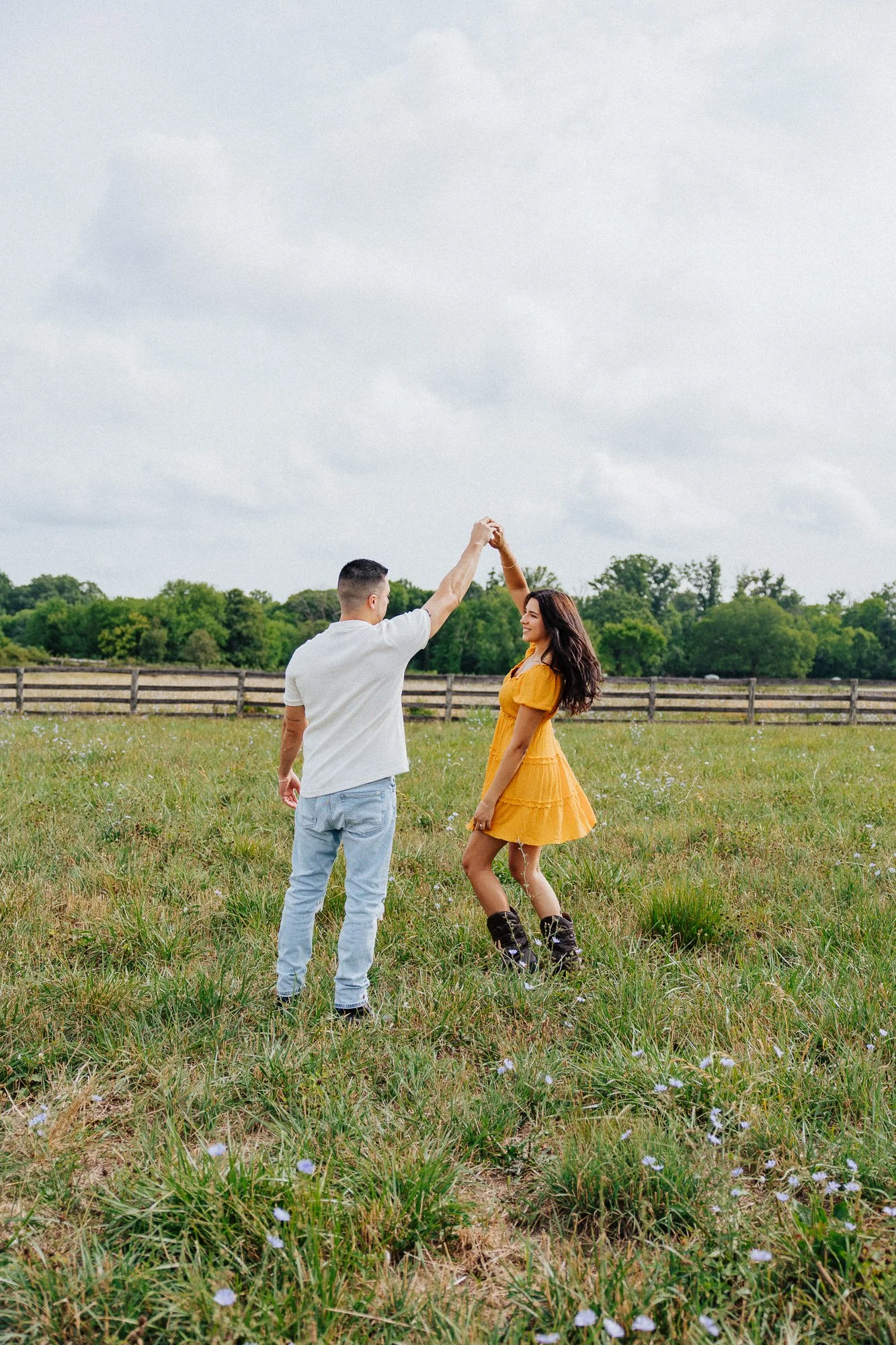 A man and woman dancing in a grassy field with a wooden fence and trees in the background, overcast sky.