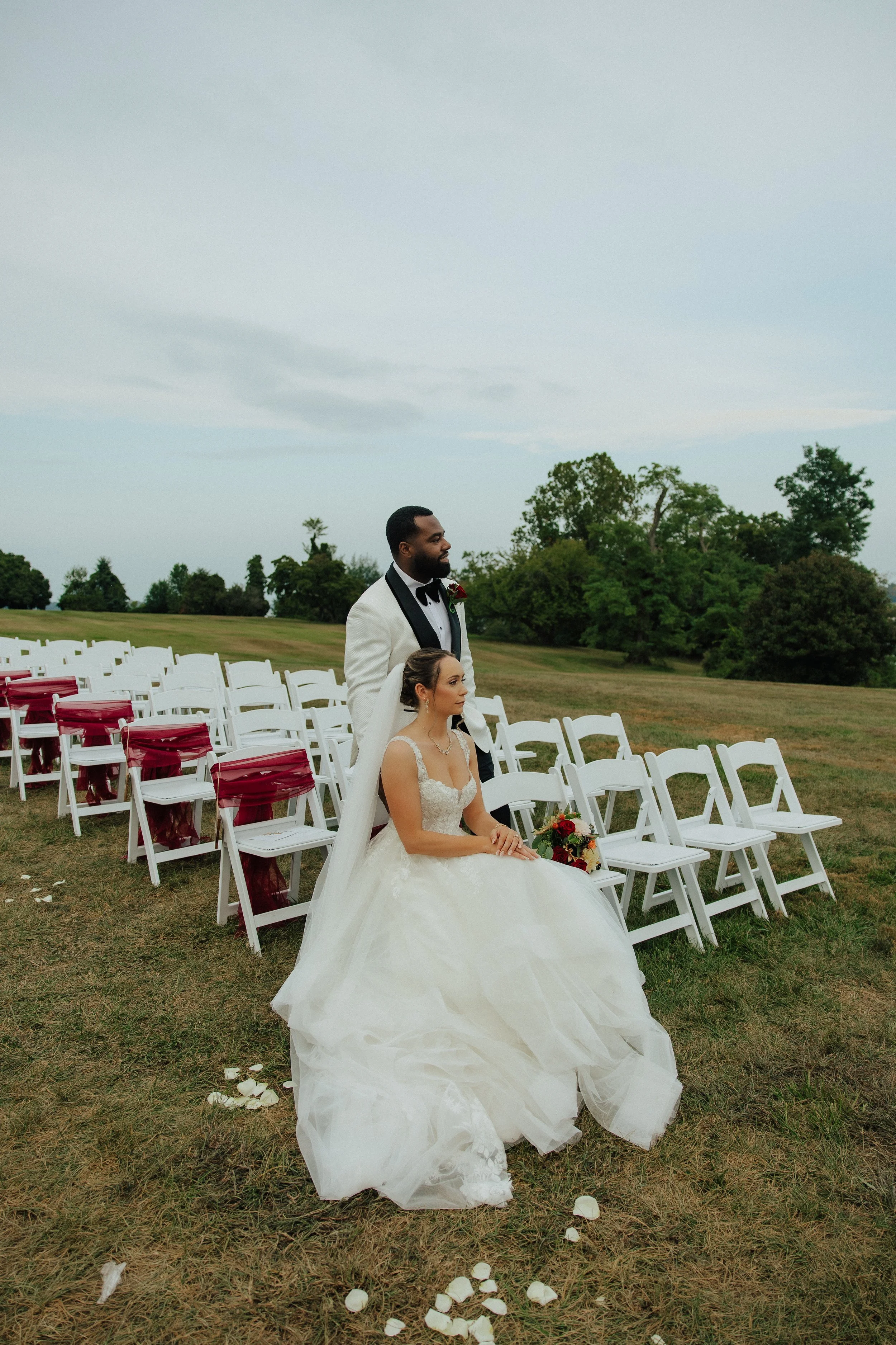 A bride in a white wedding gown sits in a chair decorated with white and burgundy cloth, with a groom standing behind her in a tuxedo outdoors during a cloudy day. There are empty chairs arranged in rows behind them, surrounded by green trees.
