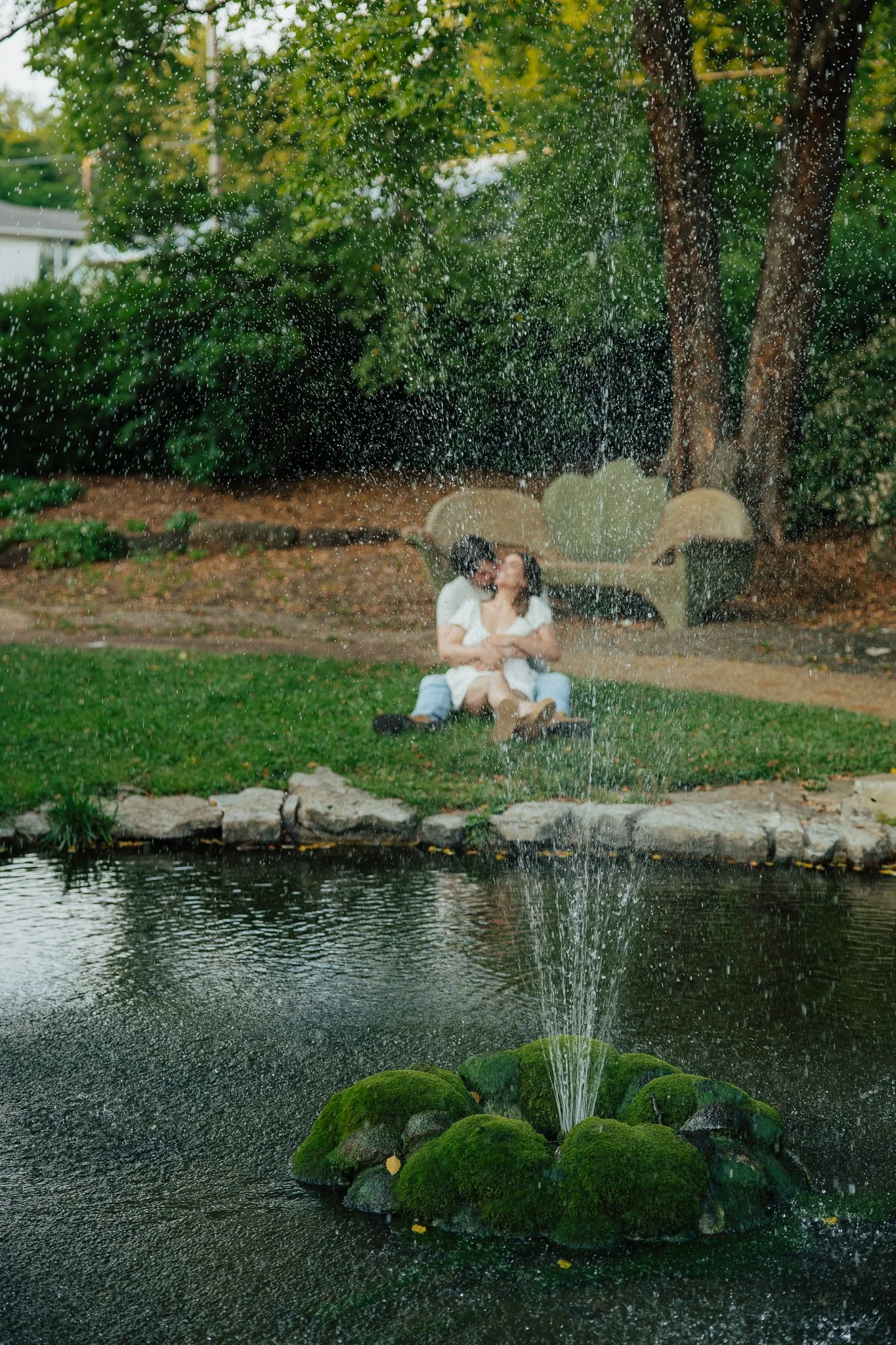 Couple sitting on a park bench near a pond with water fountain, surrounded by trees and green foliage, some leaves falling.