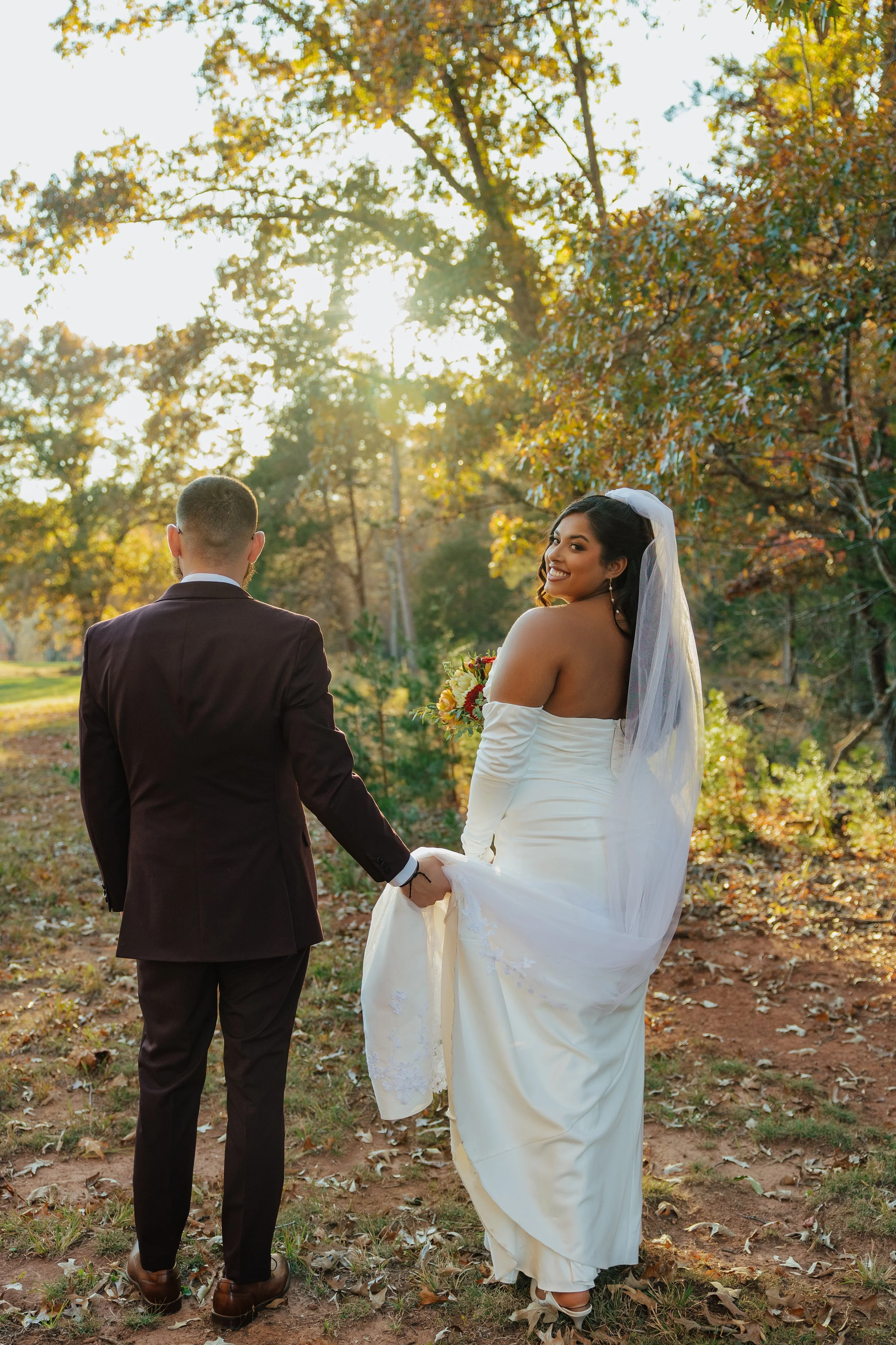 A bride in a white wedding dress and veil holding a bouquet, smiling at the camera, while a groom in a dark suit faces away, holding hands outdoors during sunset with trees and autumn leaves.