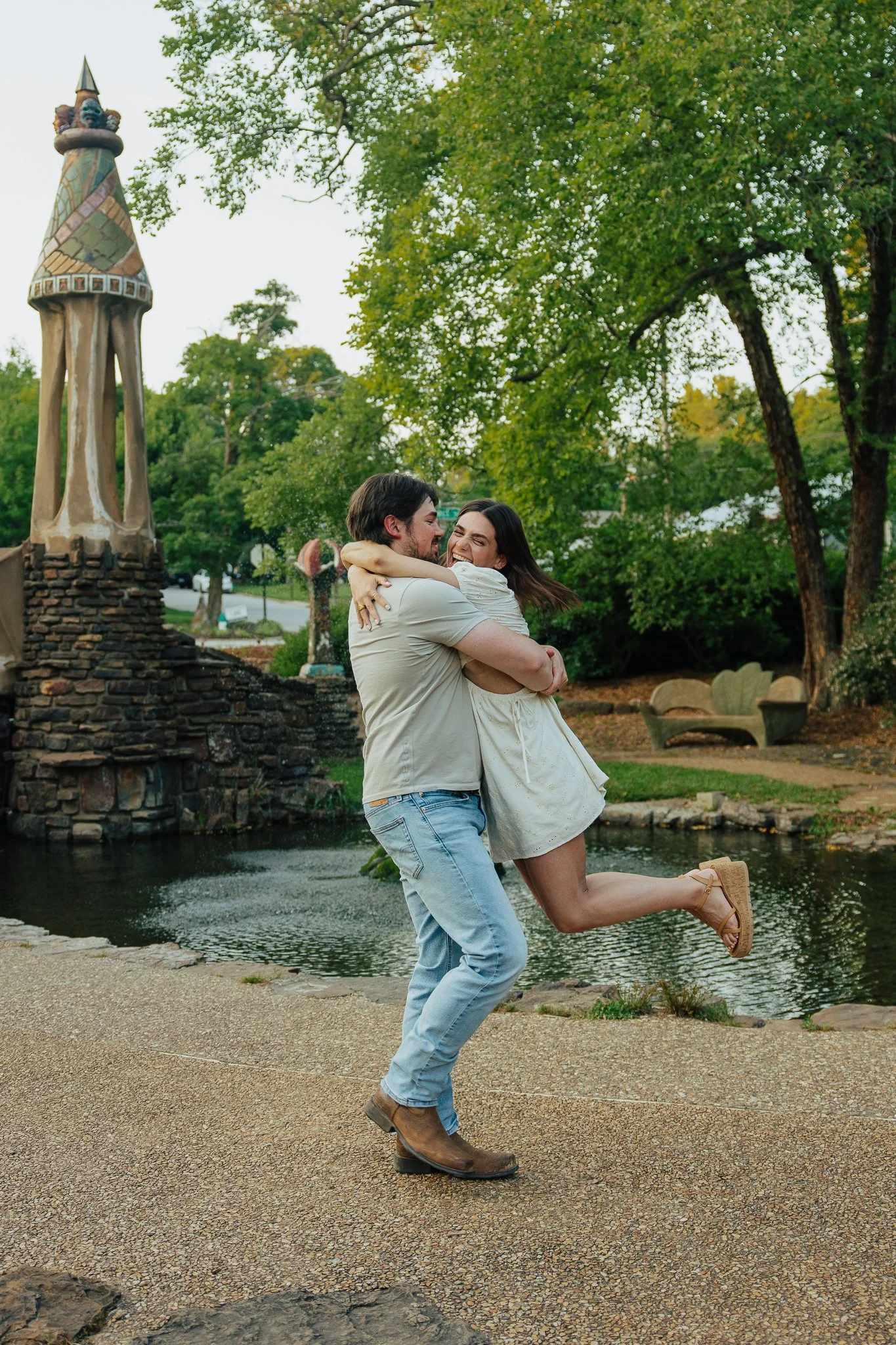 A couple hugging and smiling near a pond in a park with trees and a statue in the background.
