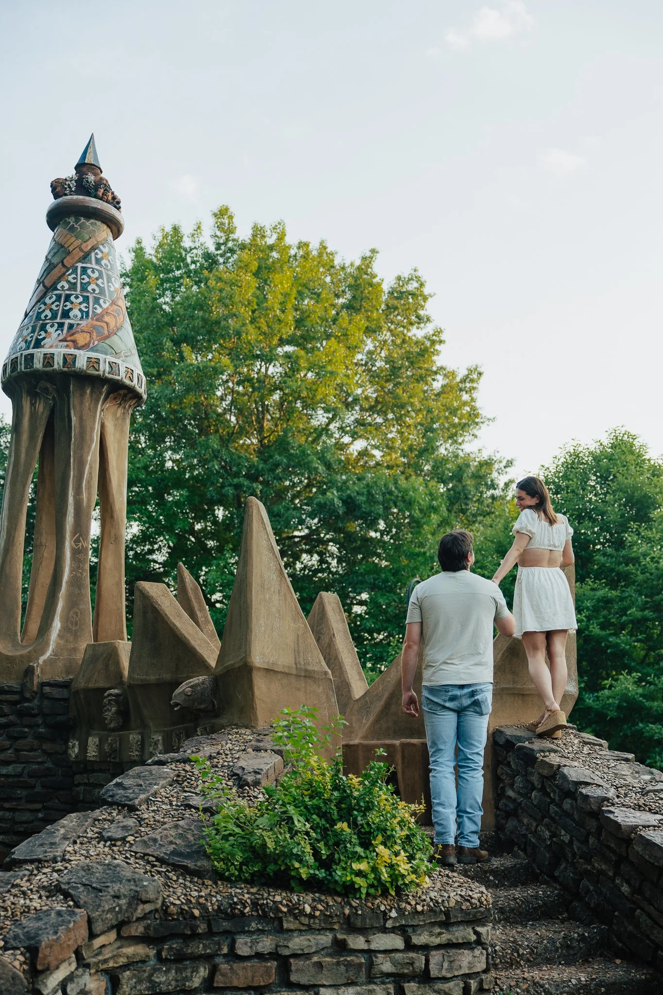 A couple walking on a rocky path in a park with large, decorative sculptures and green trees, during late afternoon or early evening.