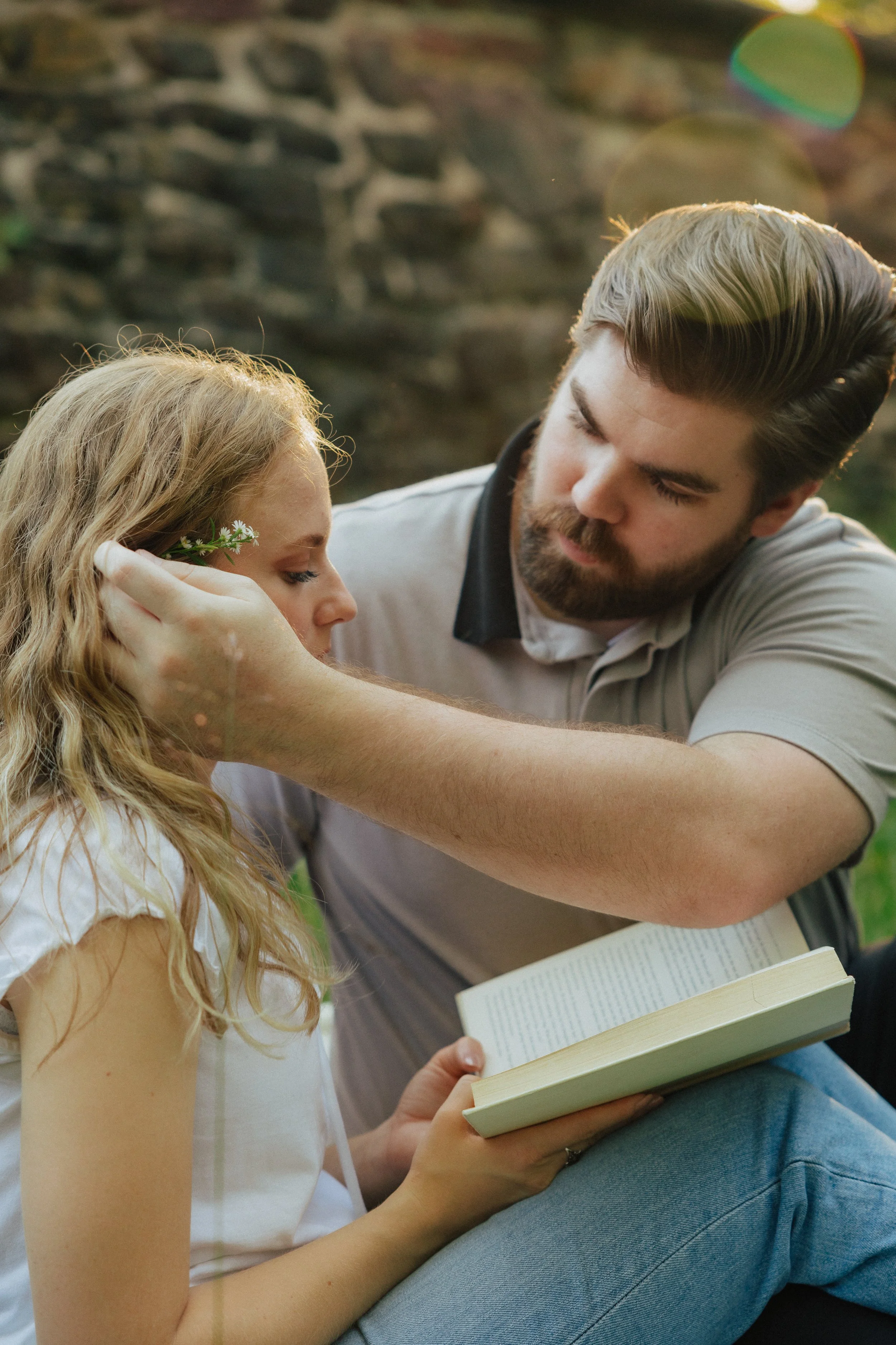 A young woman with long, wavy blonde hair and a small white flower tucked behind her ear, reading a book while sitting on a person's lap. The person, with dark hair and a beard, is also reading and has a book open on their lap. They are outdoors with