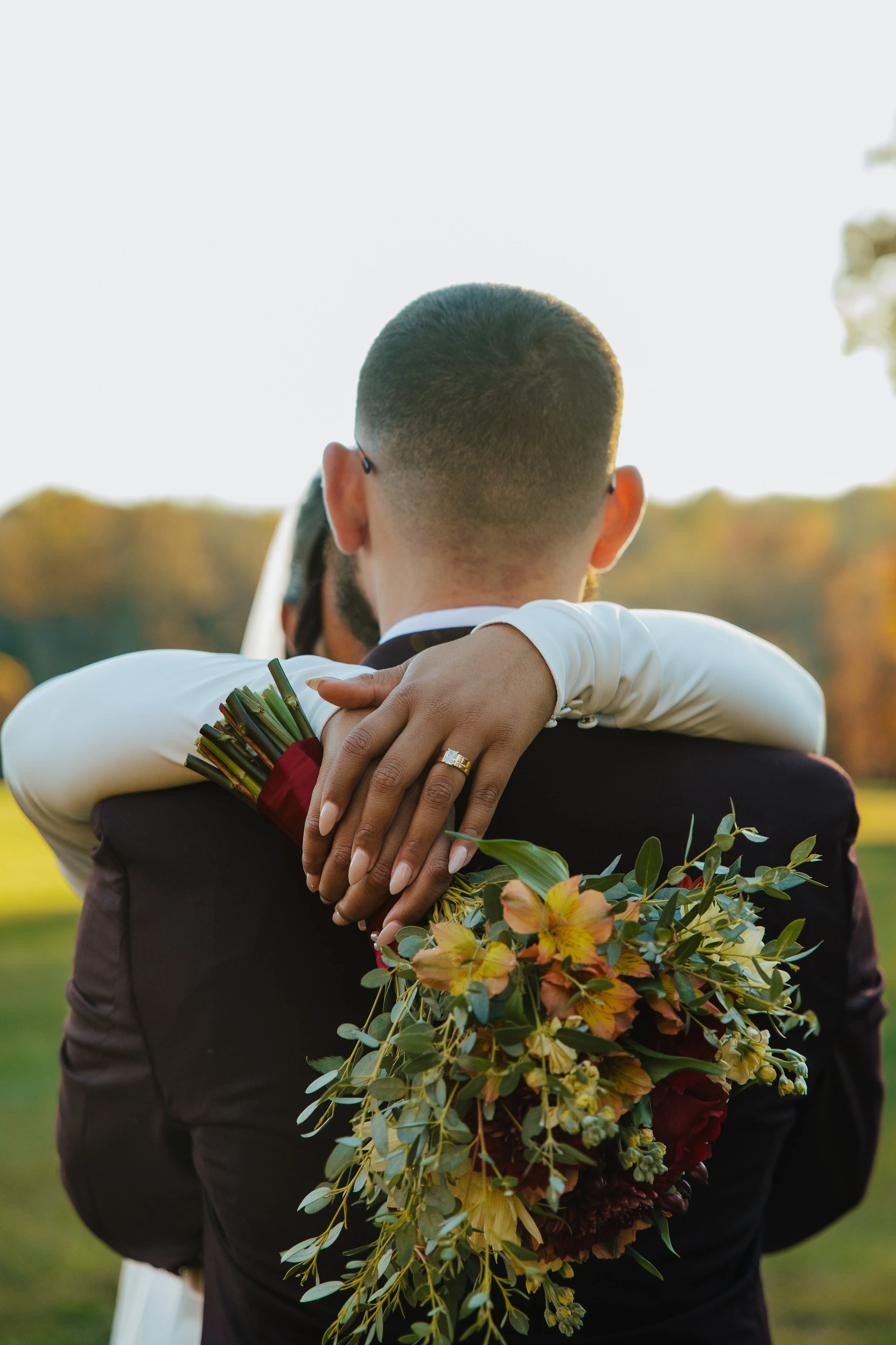 Back of a newlywed couple hugging outdoors during sunset, with the bride holding a bouquet of flowers.