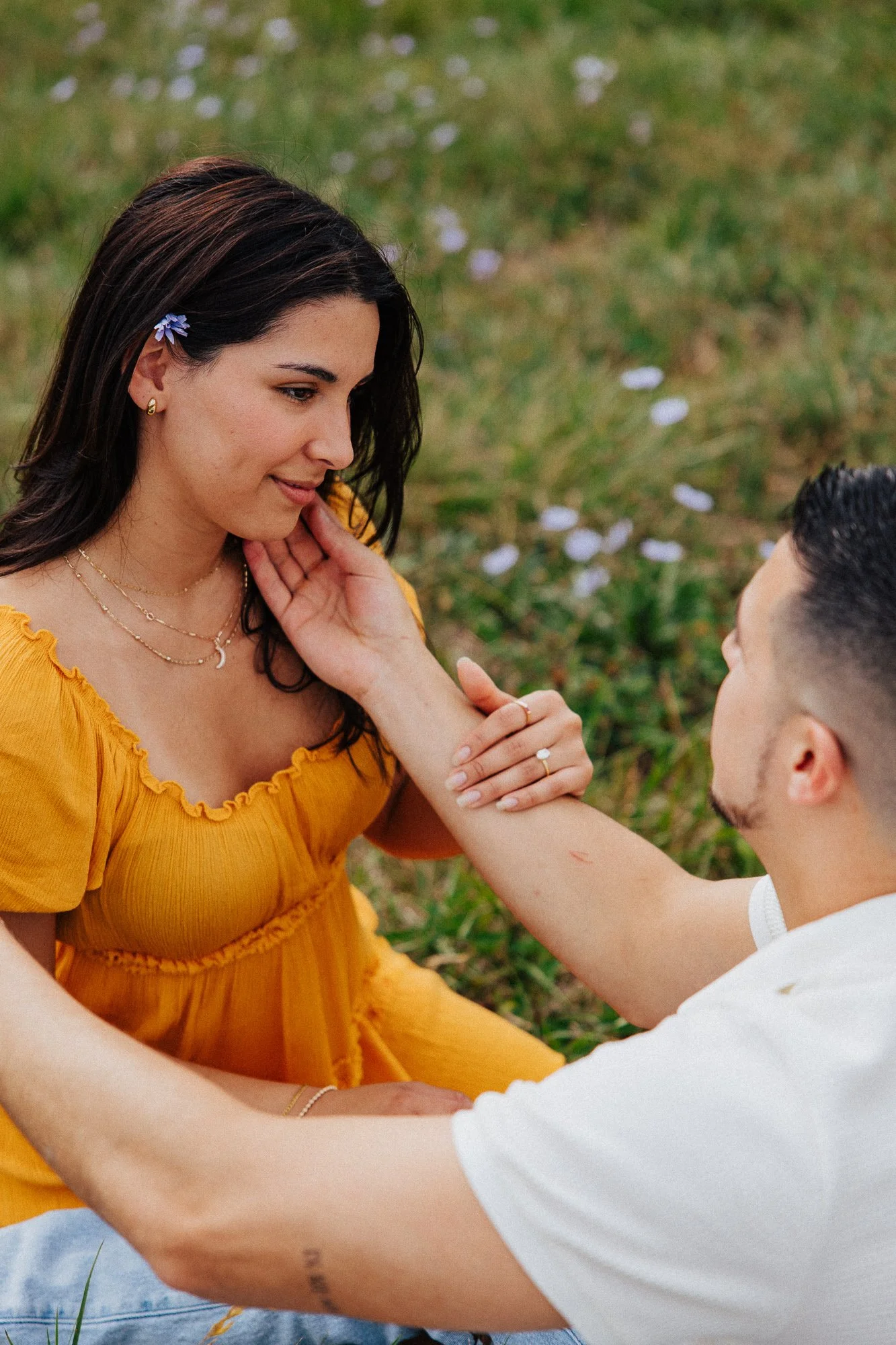 A woman in a yellow dress looking at a man, with her hand touching his face, outdoors with green grass and white flowers.