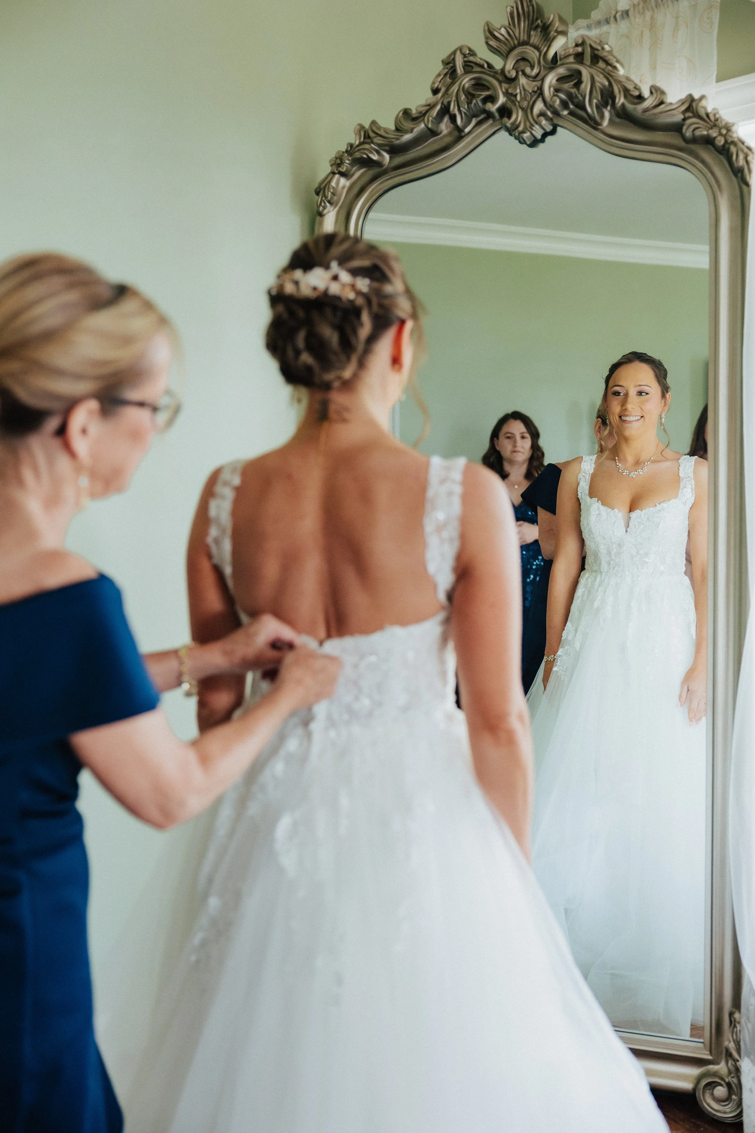 A bride in a white wedding dress smiling at her reflection in a large ornate mirror, with a woman helping her adjust her dress, and another woman observing in the background.