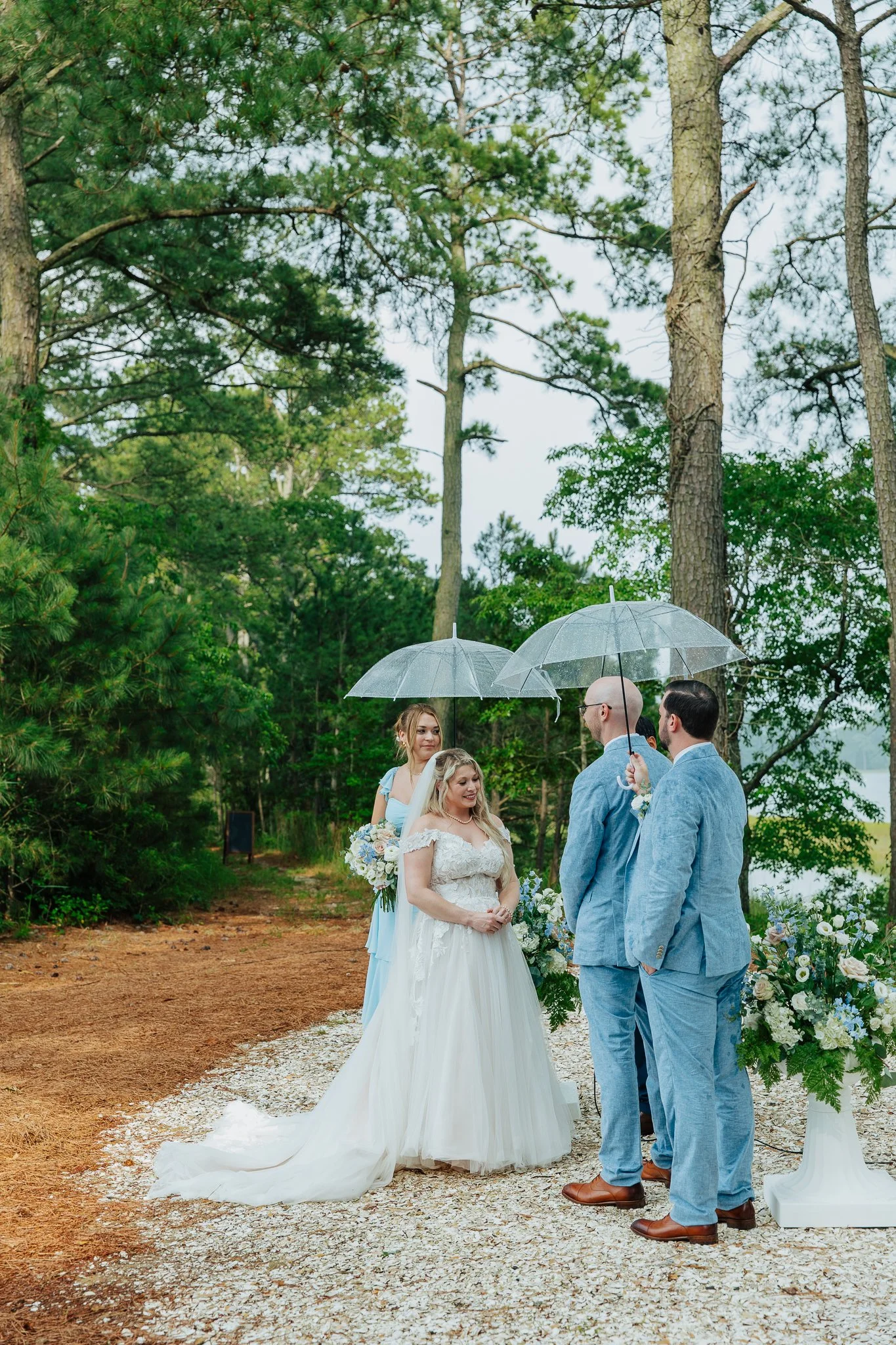A wedding ceremony outdoors with a bride and groom standing together, surrounded by bridesmaids and groomsmen. The bride is in a white gown with a long train, holding a bouquet, and the groom is in a light blue suit. The wedding party is holding umbr