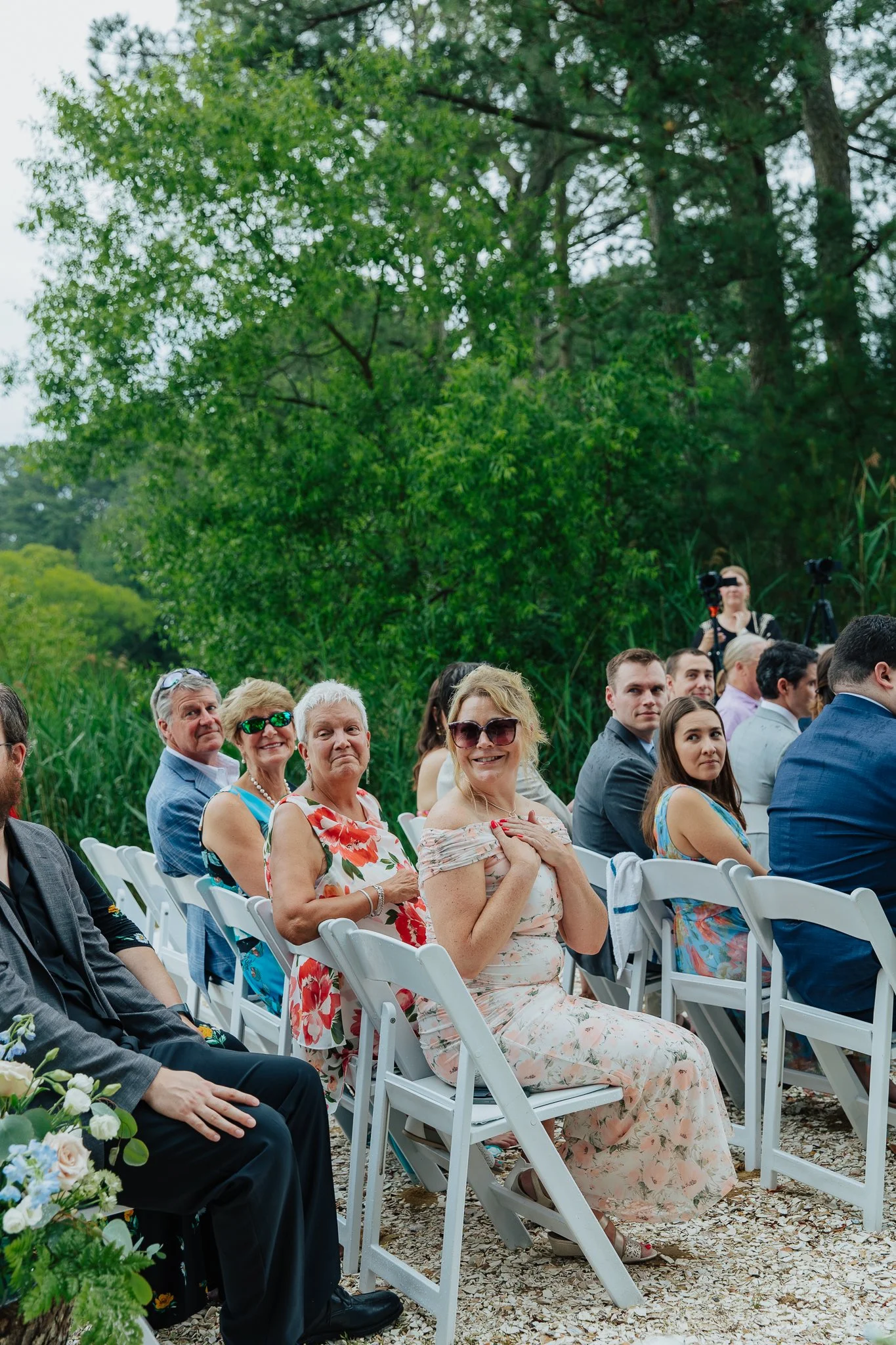 People sitting in white chairs outdoors near green trees during an event or gathering.