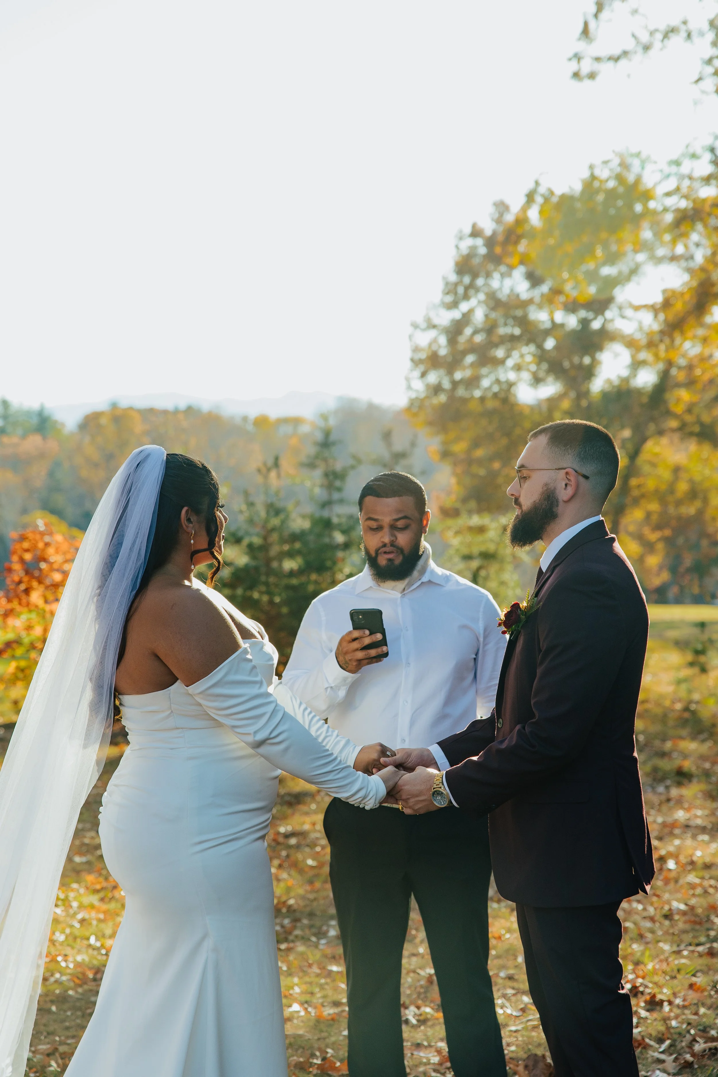 A wedding ceremony outdoors with a bride and groom holding hands, facing each other, with an officiant reading from a phone. The setting has autumn foliage and warm fall colors.