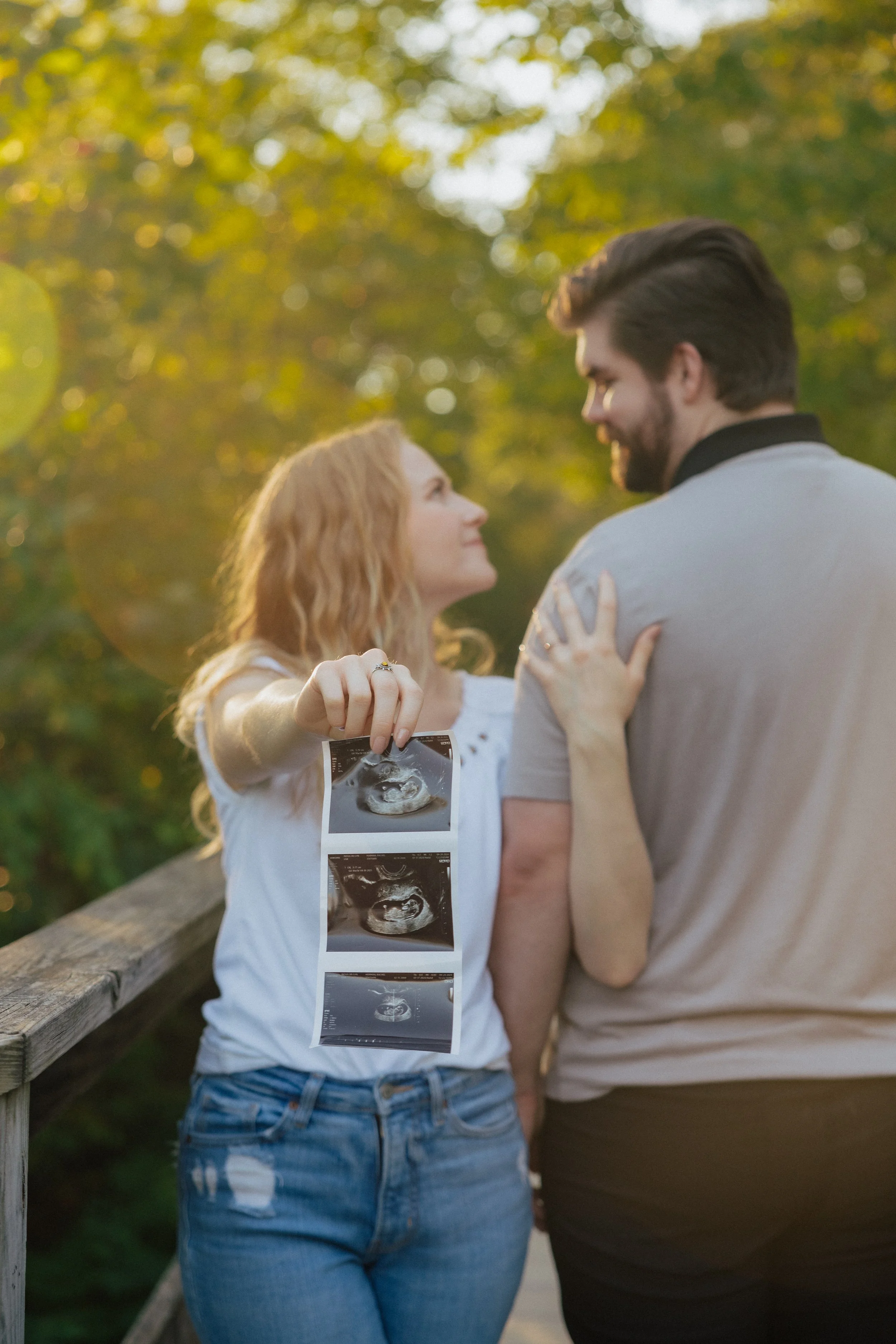A pregnant woman and a man standing outdoors on a wooden bridge, the woman holding ultrasound images of her pregnancy, with trees and sunlight in the background.