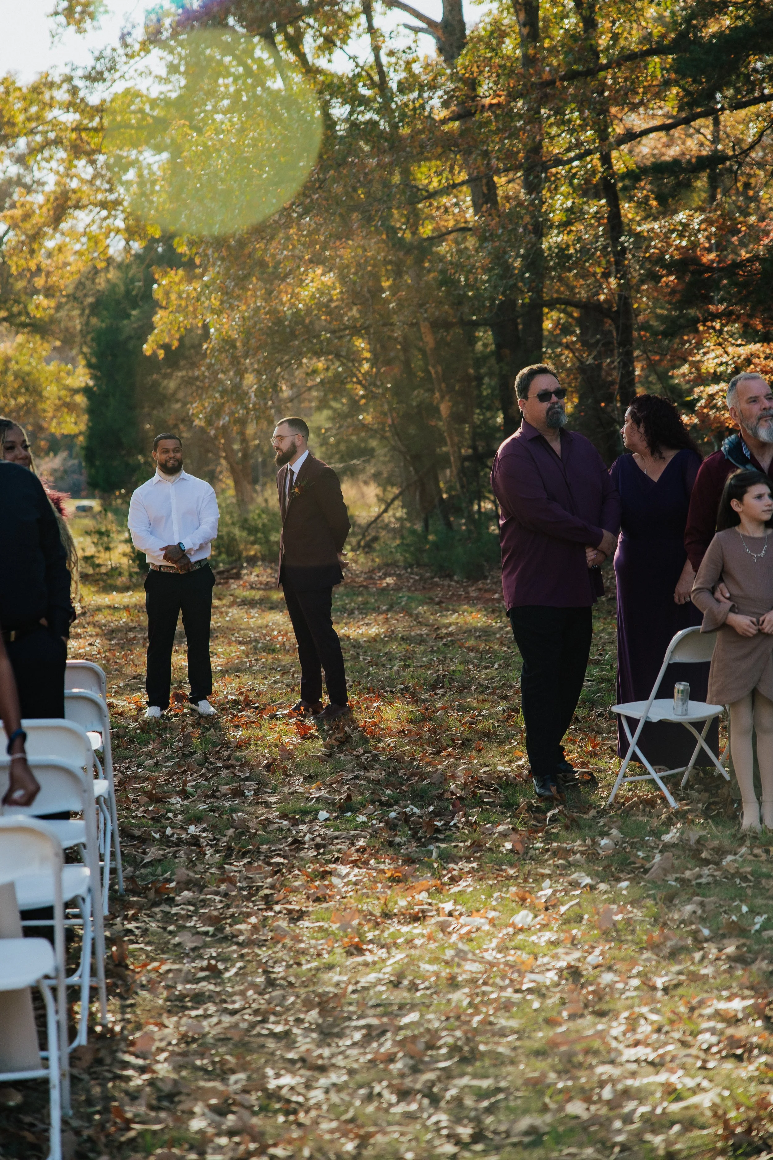 People gathered outdoors in a park during autumn, with colorful leaves on the ground and trees. Some individuals are standing and conversing while others are seated on white chairs, suggesting a social or celebratory event.
