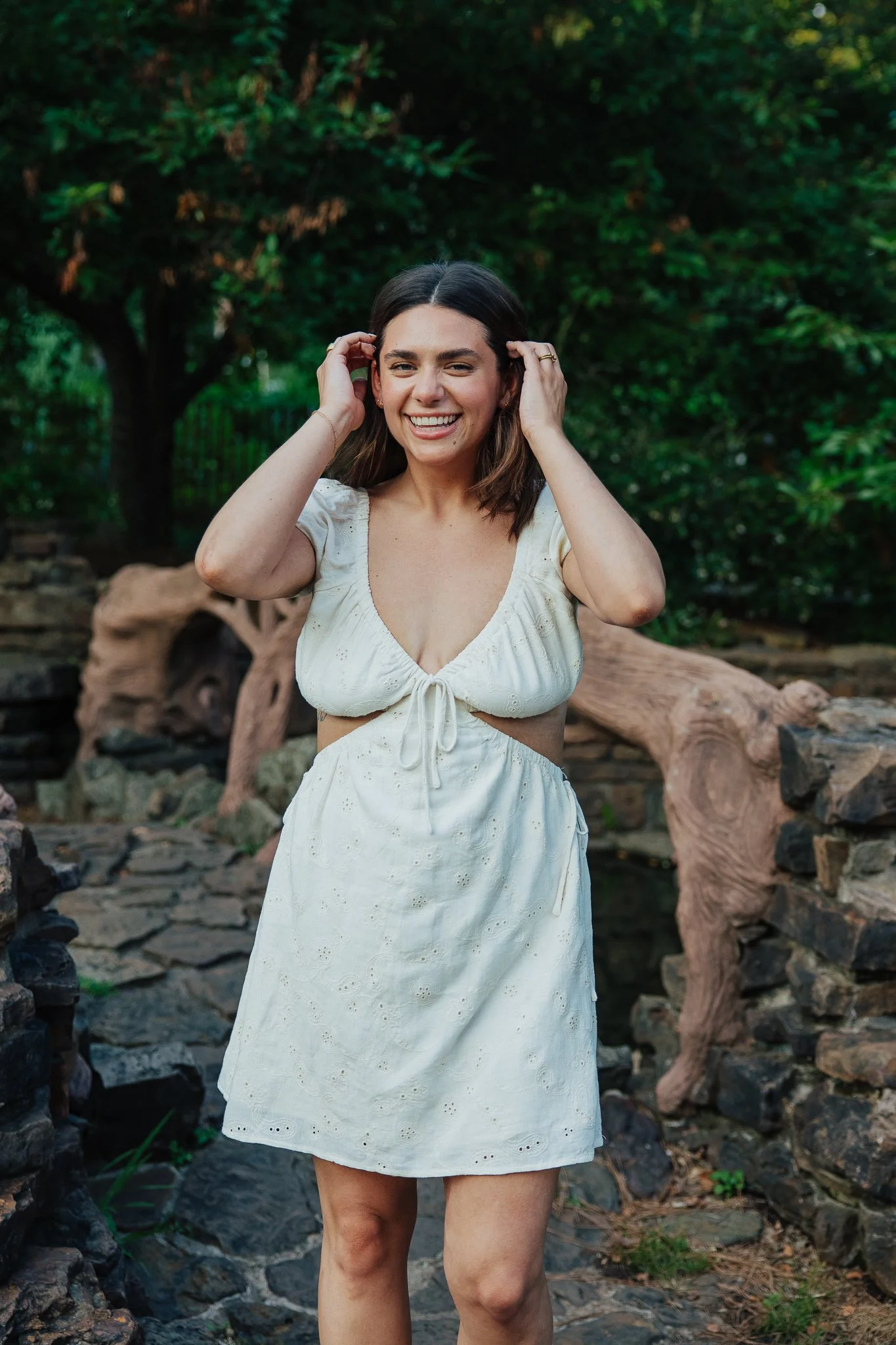 A woman with shoulder-length brown hair, wearing a white dress with eyelet details, standing outdoors near rocks and a large twisted tree trunk, with green foliage in the background. She is smiling and adjusting her hair with both hands.