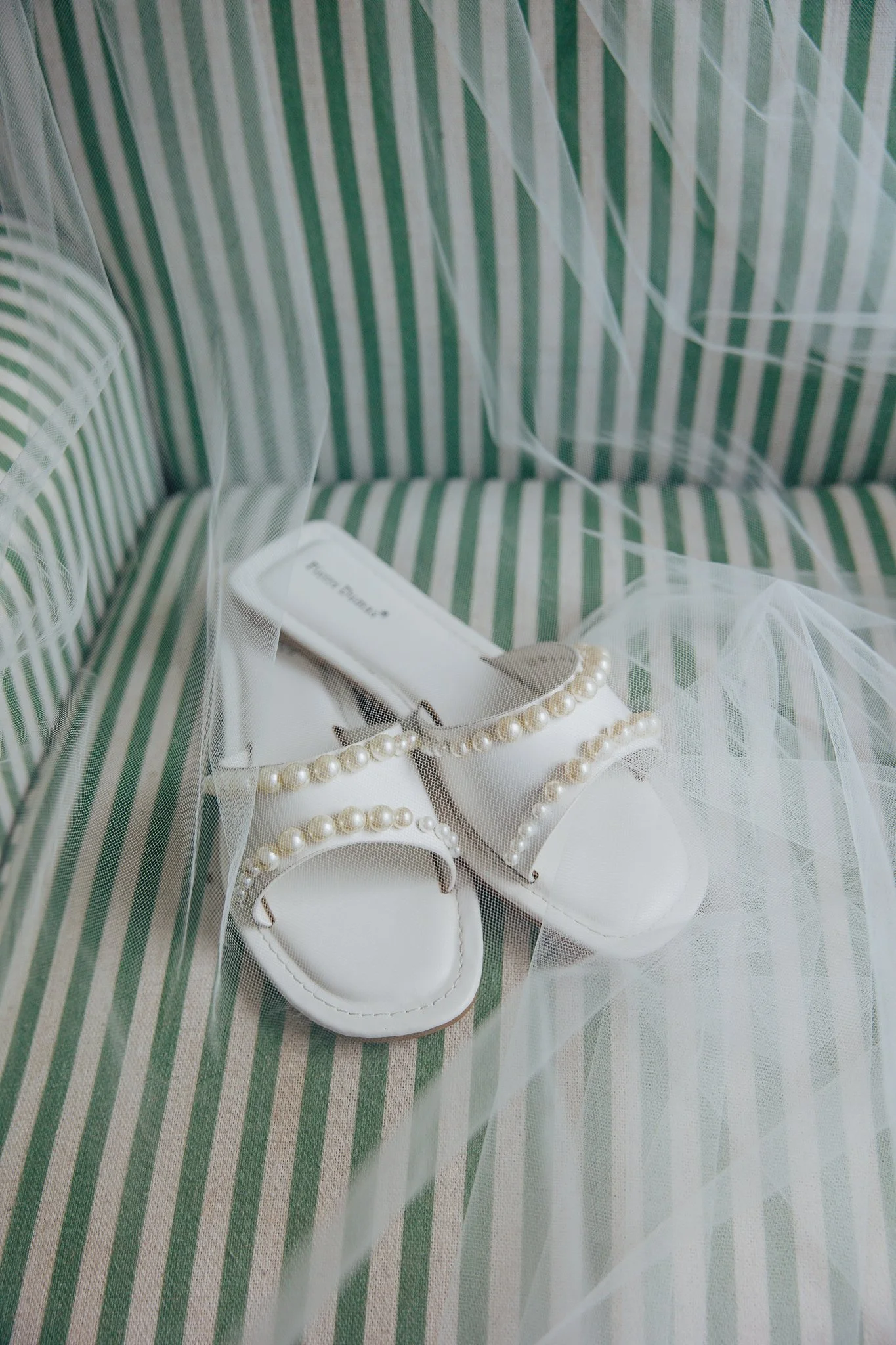 White shoes with pearl embellishments and a pearl necklace on striped fabric with green, white, and beige colors, covered by sheer white tulle.