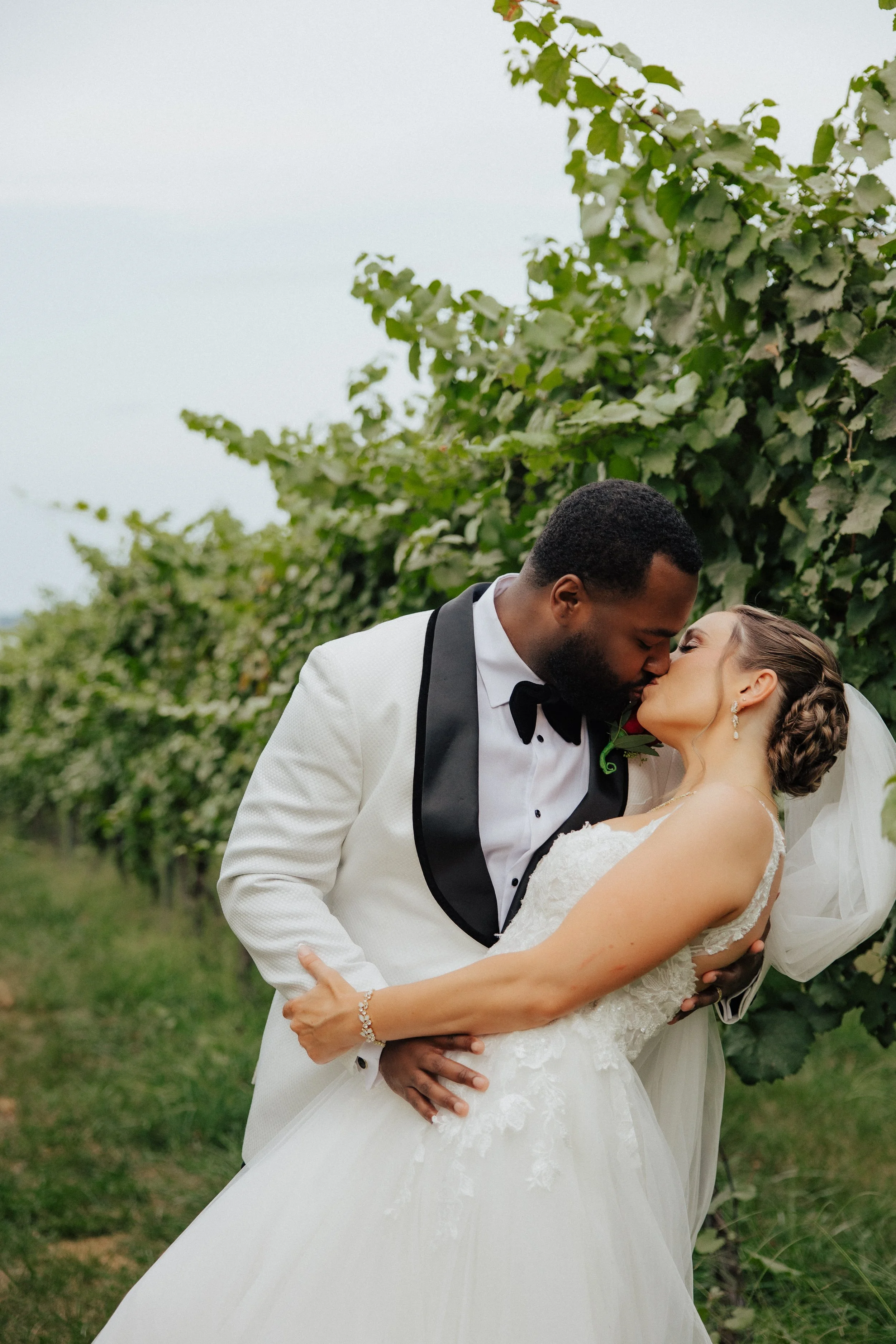 A bride and groom sharing a kiss in a vineyard, with lush green grapevines in the background.
