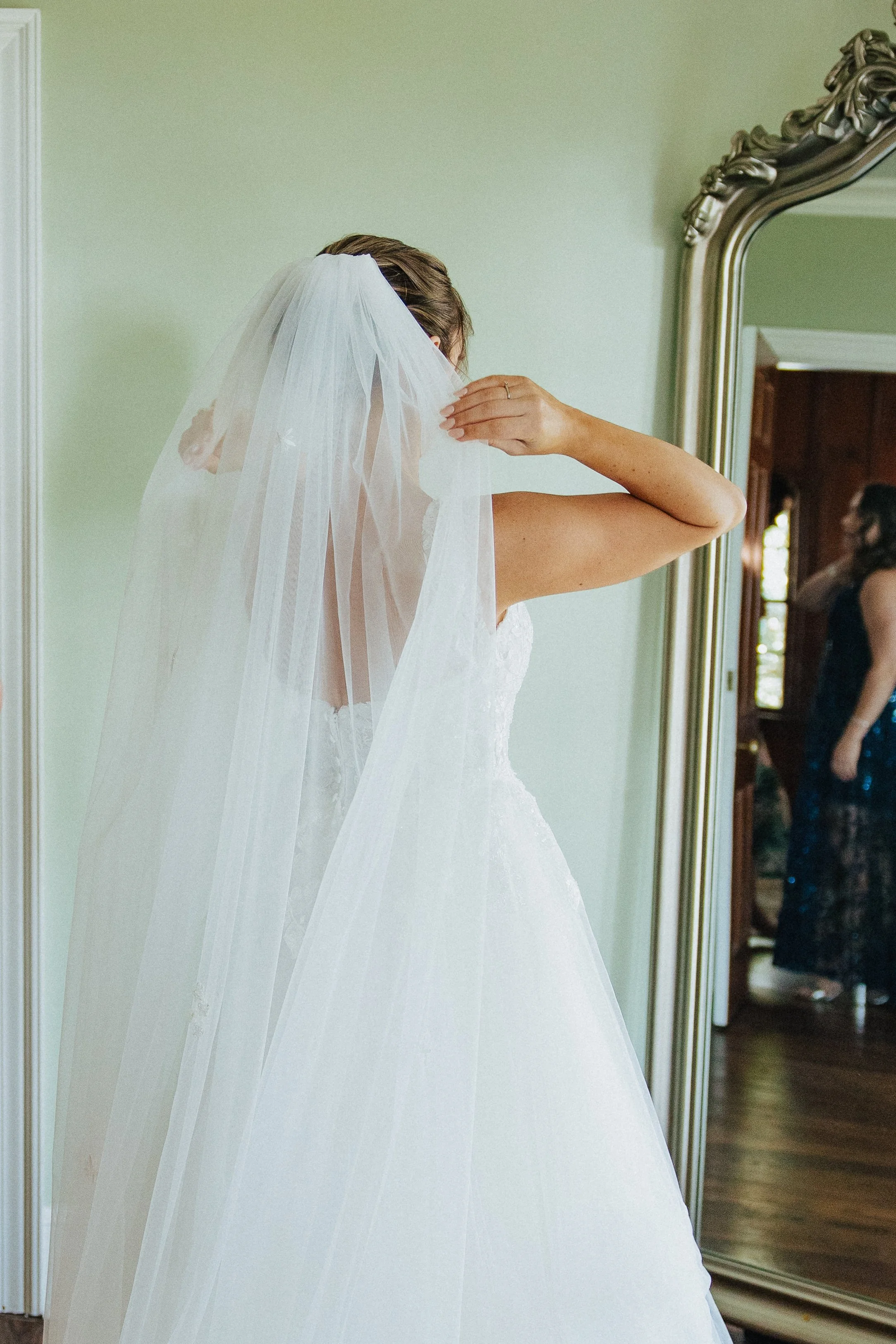 A bride adjusting her veil in front of a mirror in a room with wooden floors and green walls.