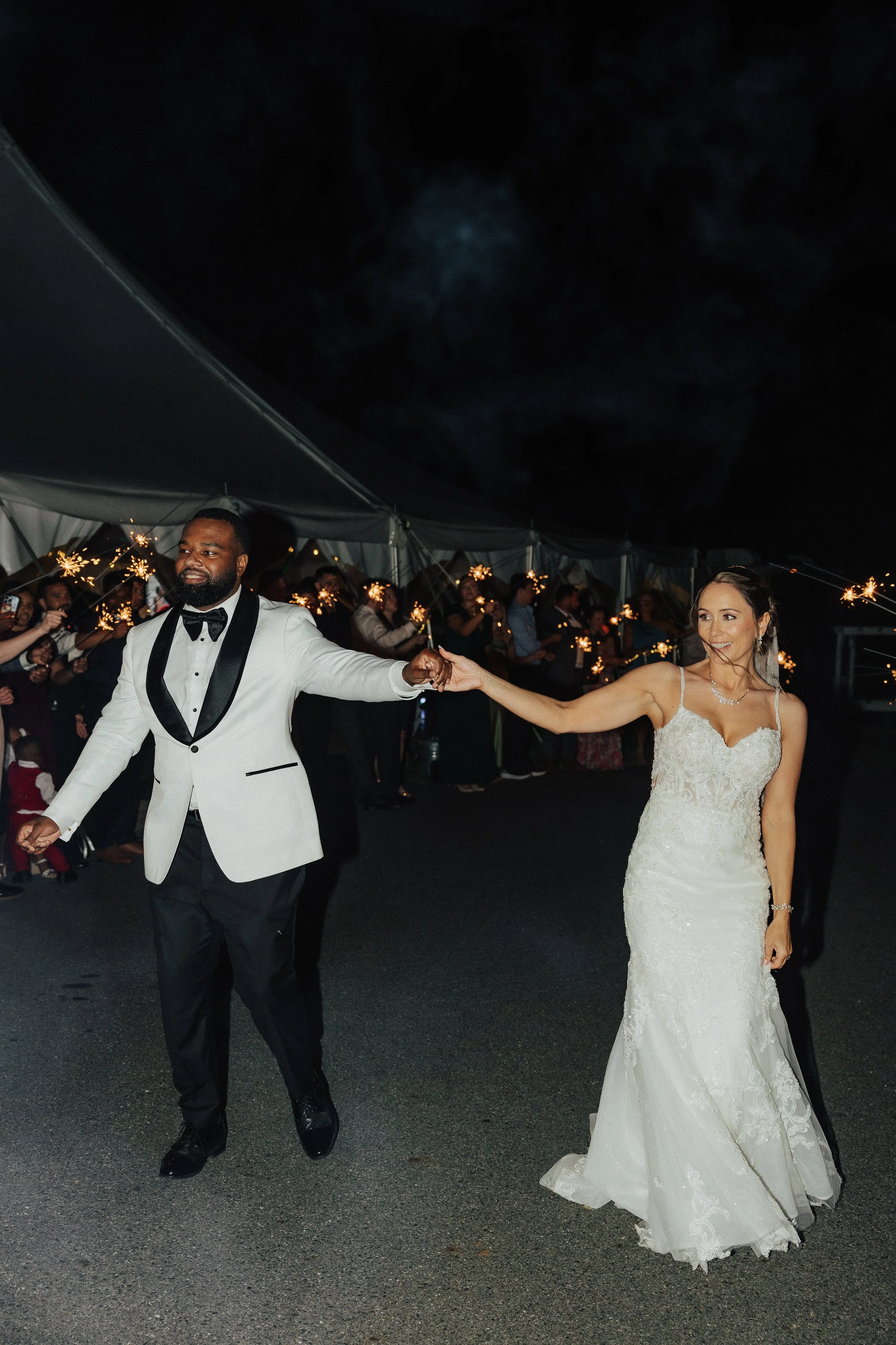 A bride and groom dancing at their wedding reception, surrounded by guests holding sparklers inside a large tent at night.