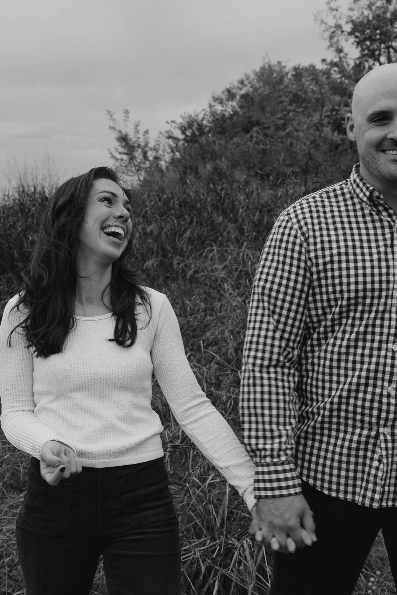 Two people holding hands and smiling, walking outdoors in a natural setting with trees and grass in the background.