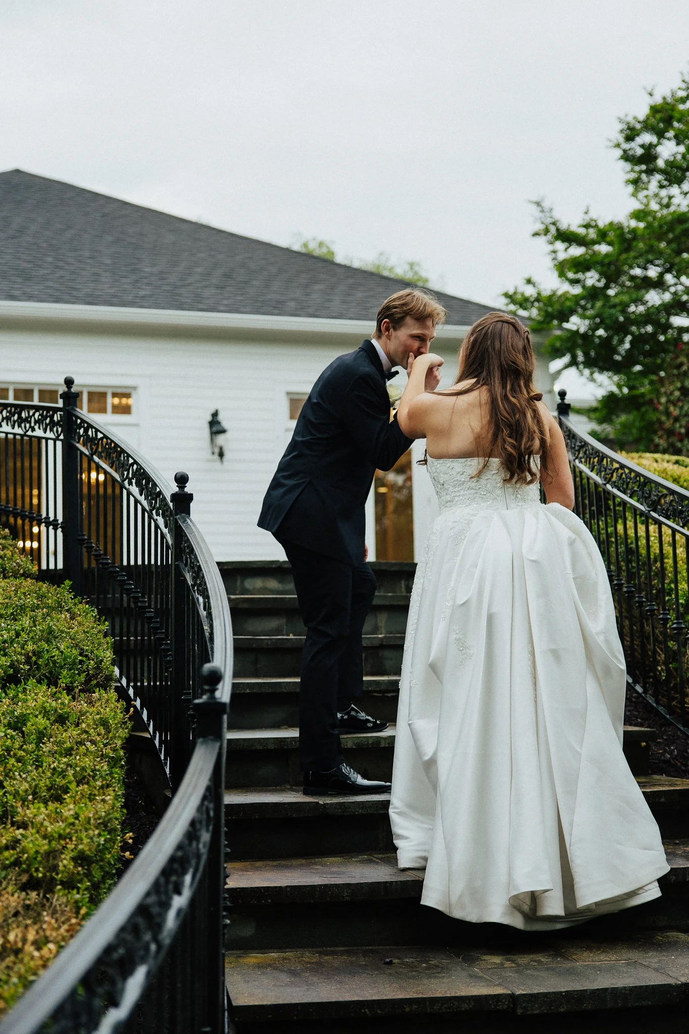 A groom kissing a bride on the cheek as they stand on outdoor steps near a white house with a black iron railing. The groom wears a dark suit, and the bride wears a white wedding gown with a long skirt. There are trees and bushes in the background on a cloudy day.