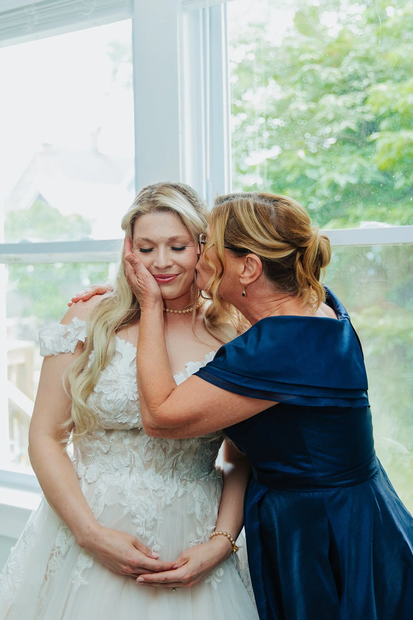 A woman in a white wedding dress with lace details has her eyes closed and a gentle smile as another woman in a navy blue dress kisses her on the cheek, with her hand tenderly touching the bride's face. The background shows a large window with green 