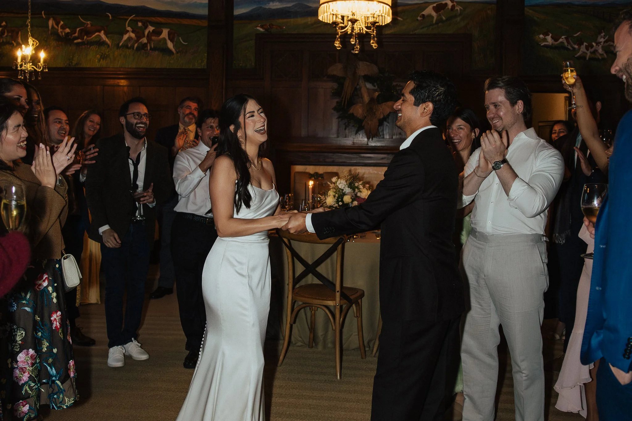 A bride and groom holding hands and smiling at each other during their wedding reception, surrounded by friends who are laughing and celebrating.