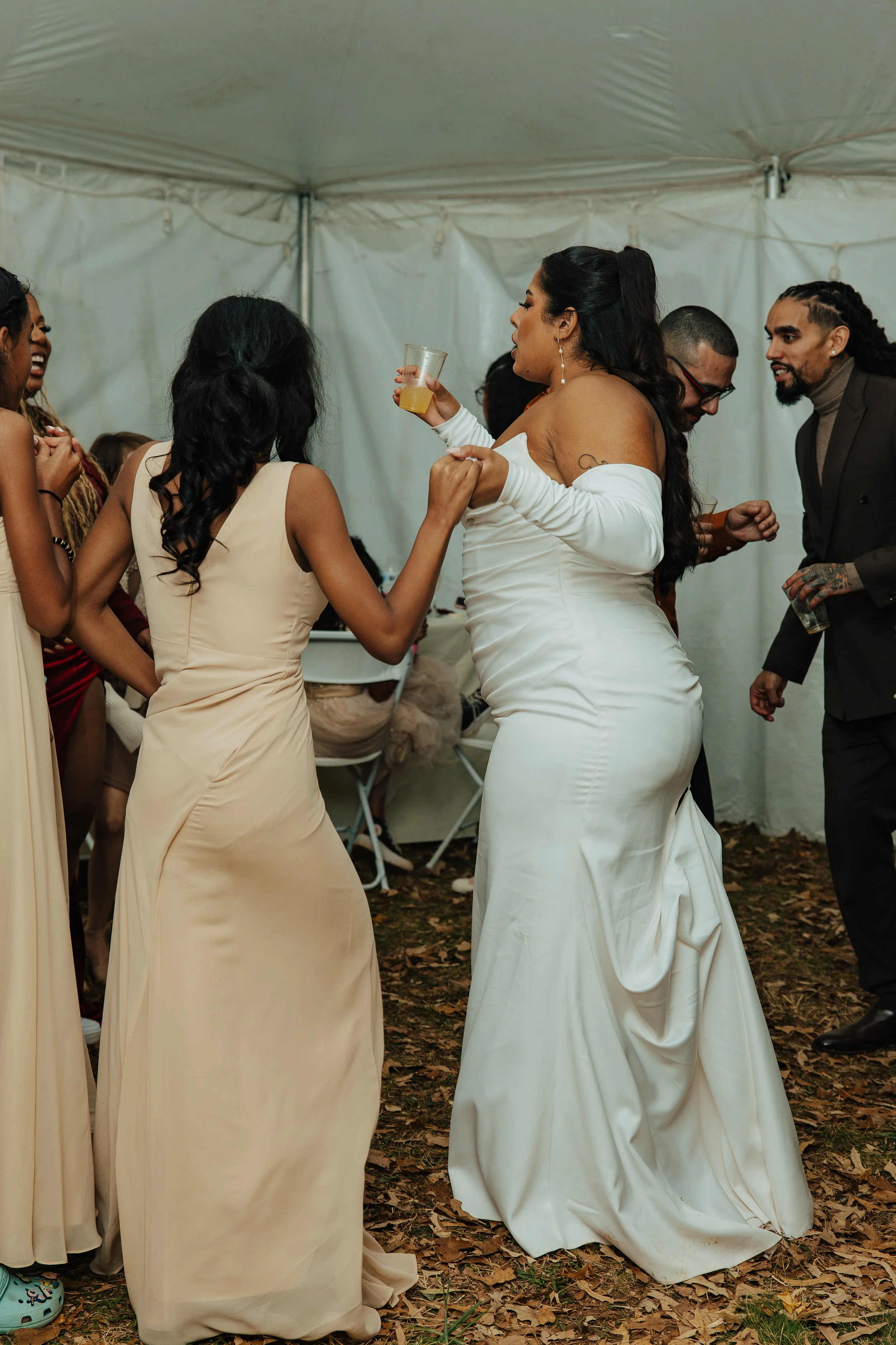 Group of women in wedding dresses and men in suits dancing at a wedding inside a tent with a leaf-covered ground.