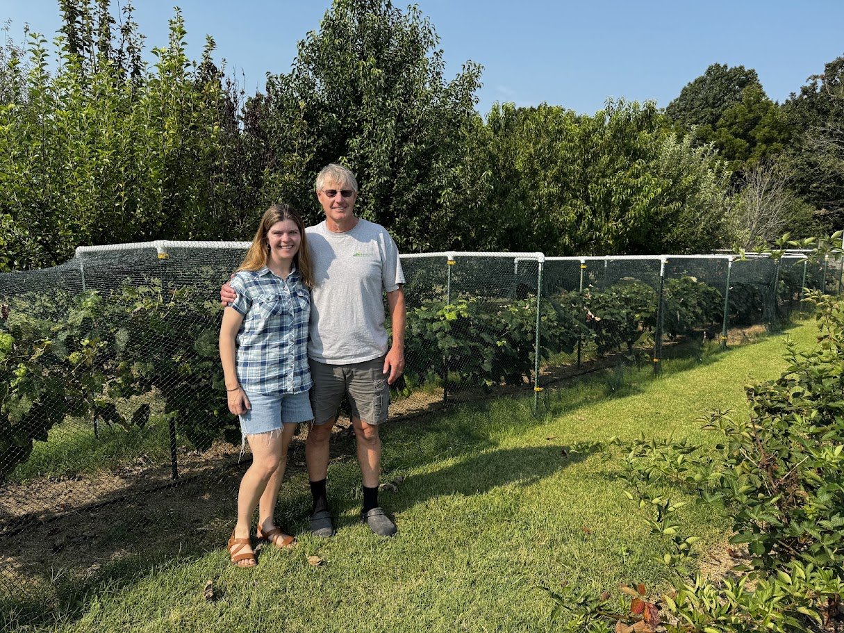 Two people standing in front of a garden with plants protected by netting, outdoors on a sunny day.
